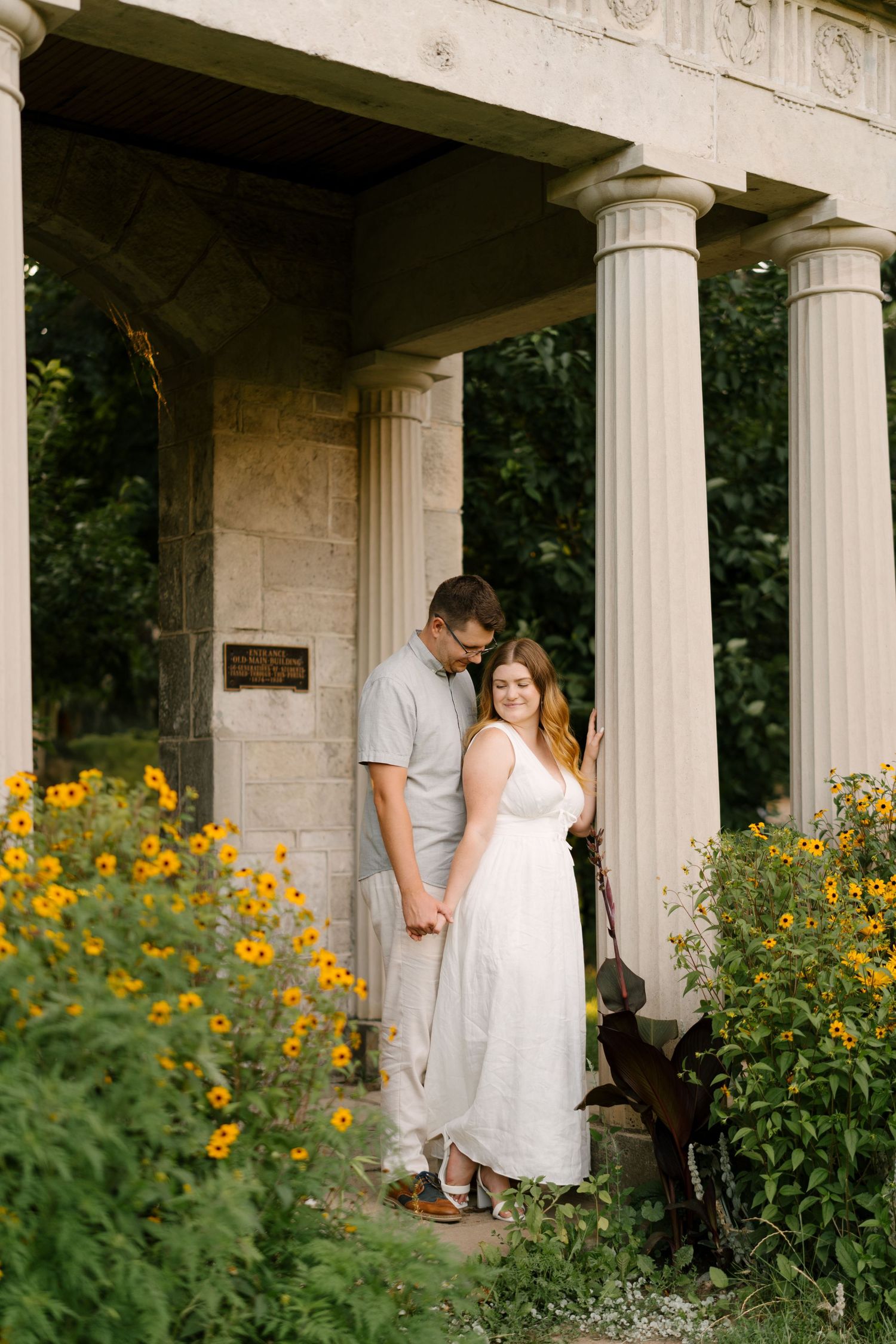 Couple holding hands under stone columns surrounded by vibrant yellow flowers at golden hour.