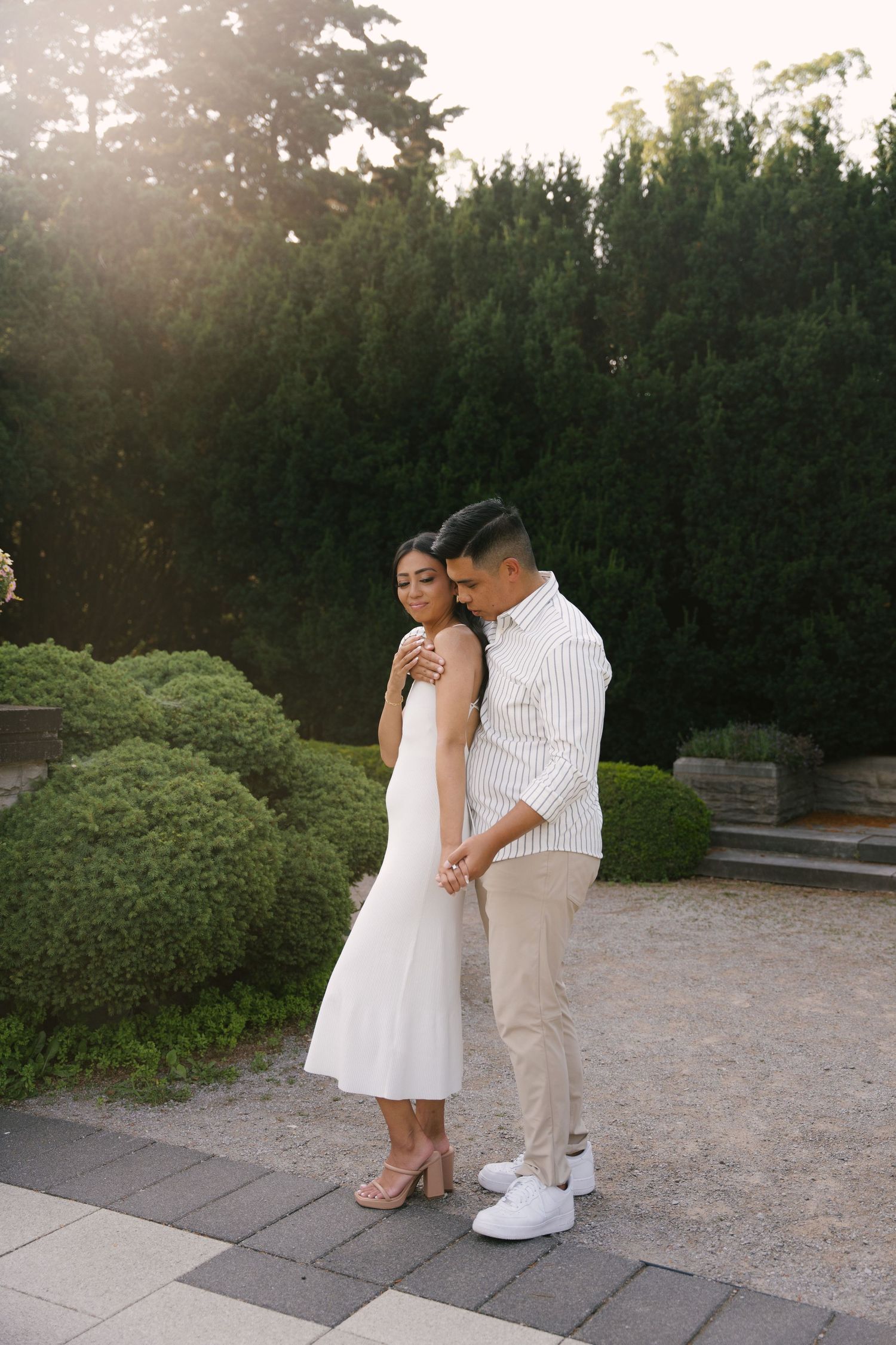 Man embracing woman in white dress from behind in a sunlit garden with manicured shrubs.