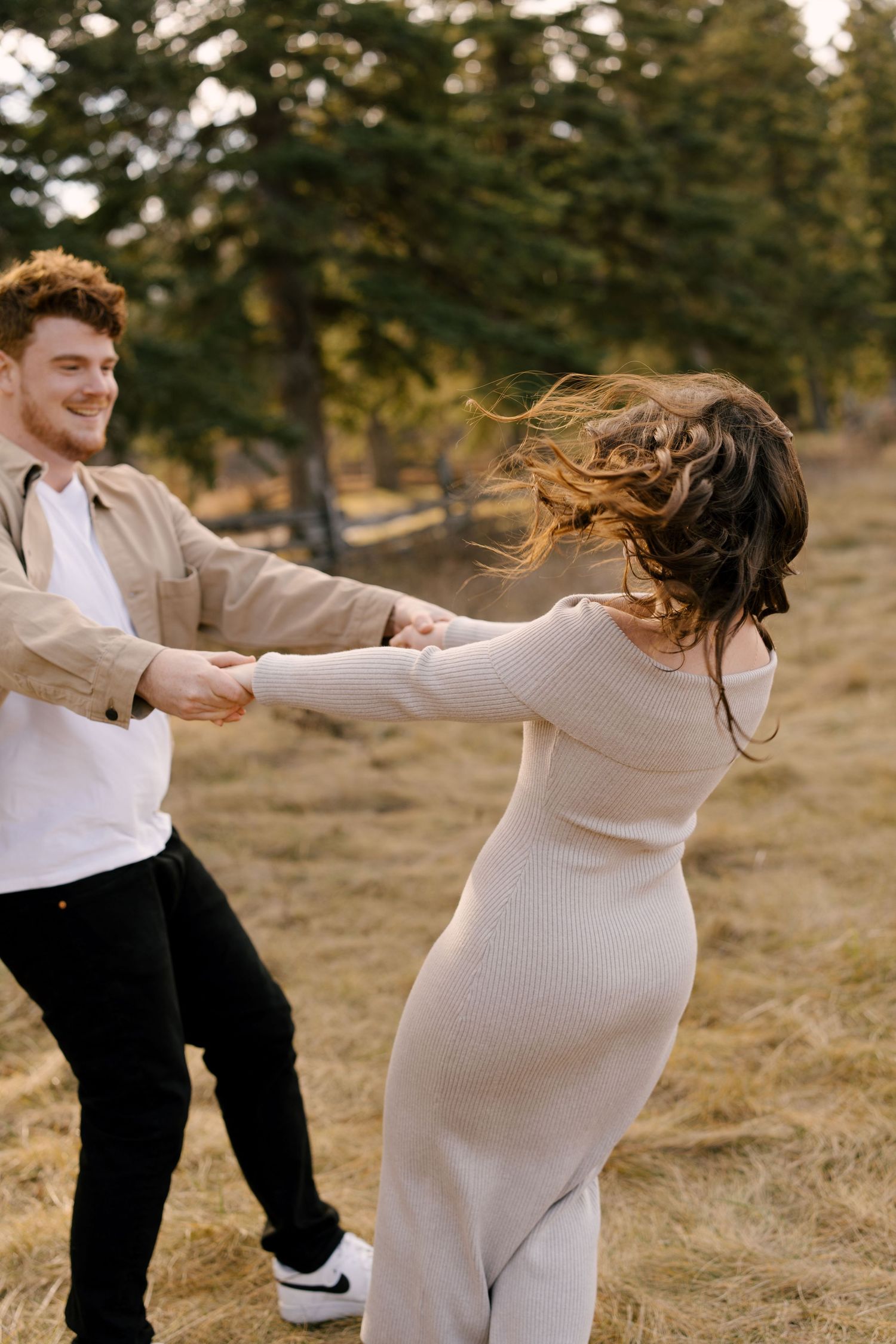 Joyful couple holding hands and spinning in a dry grass field surrounded by pine trees.