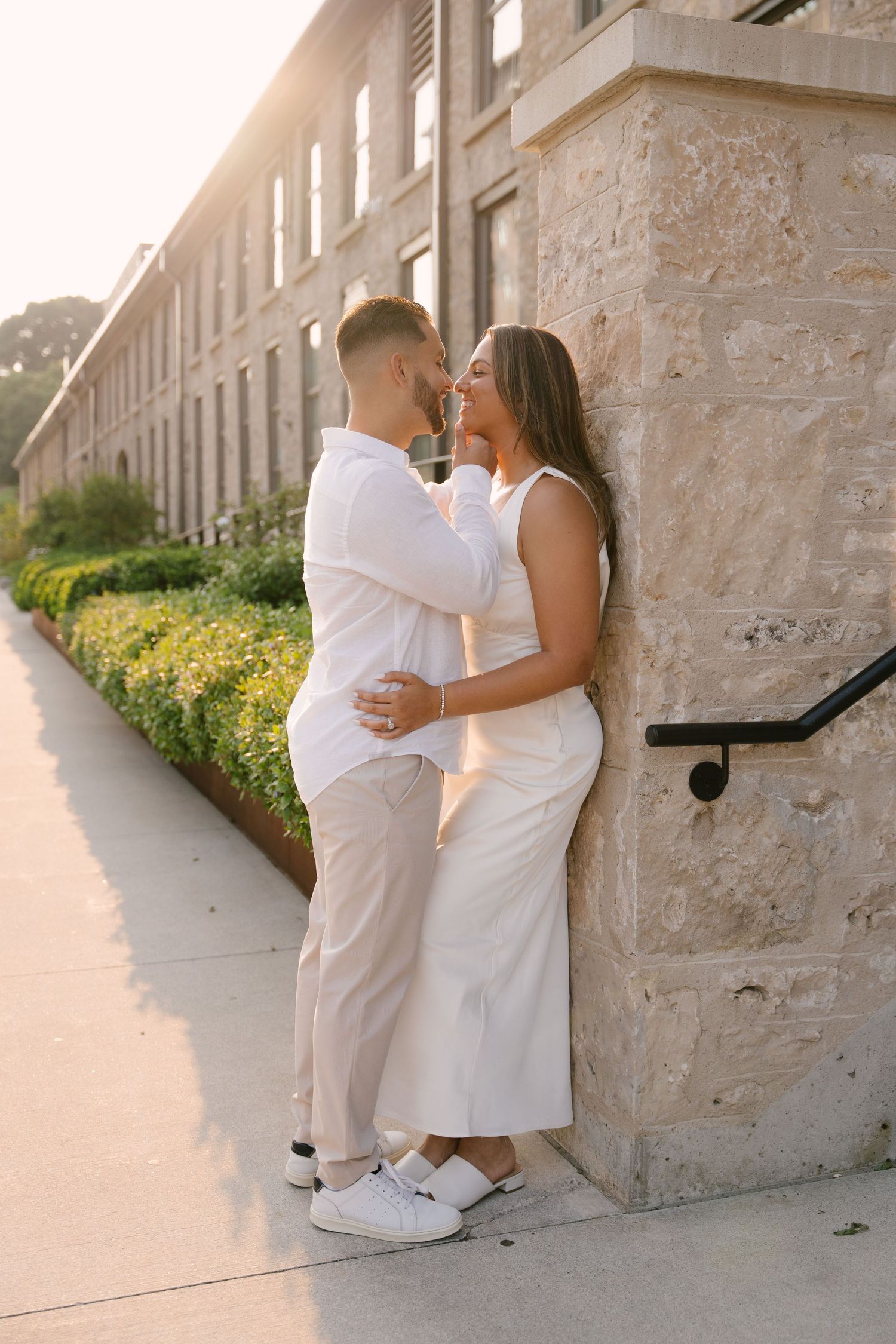 A couple in white outfits shares a romantic almost-kiss moment leaning against a stone building at sunset.