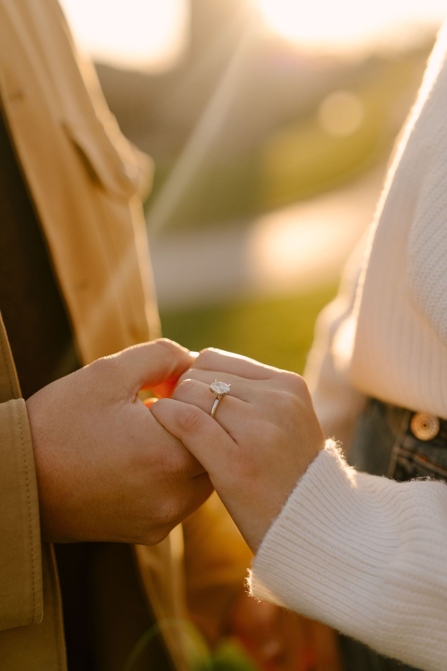 Close-up of couple holding hands at sunset revealing a diamond solitaire engagement ring on her finger.