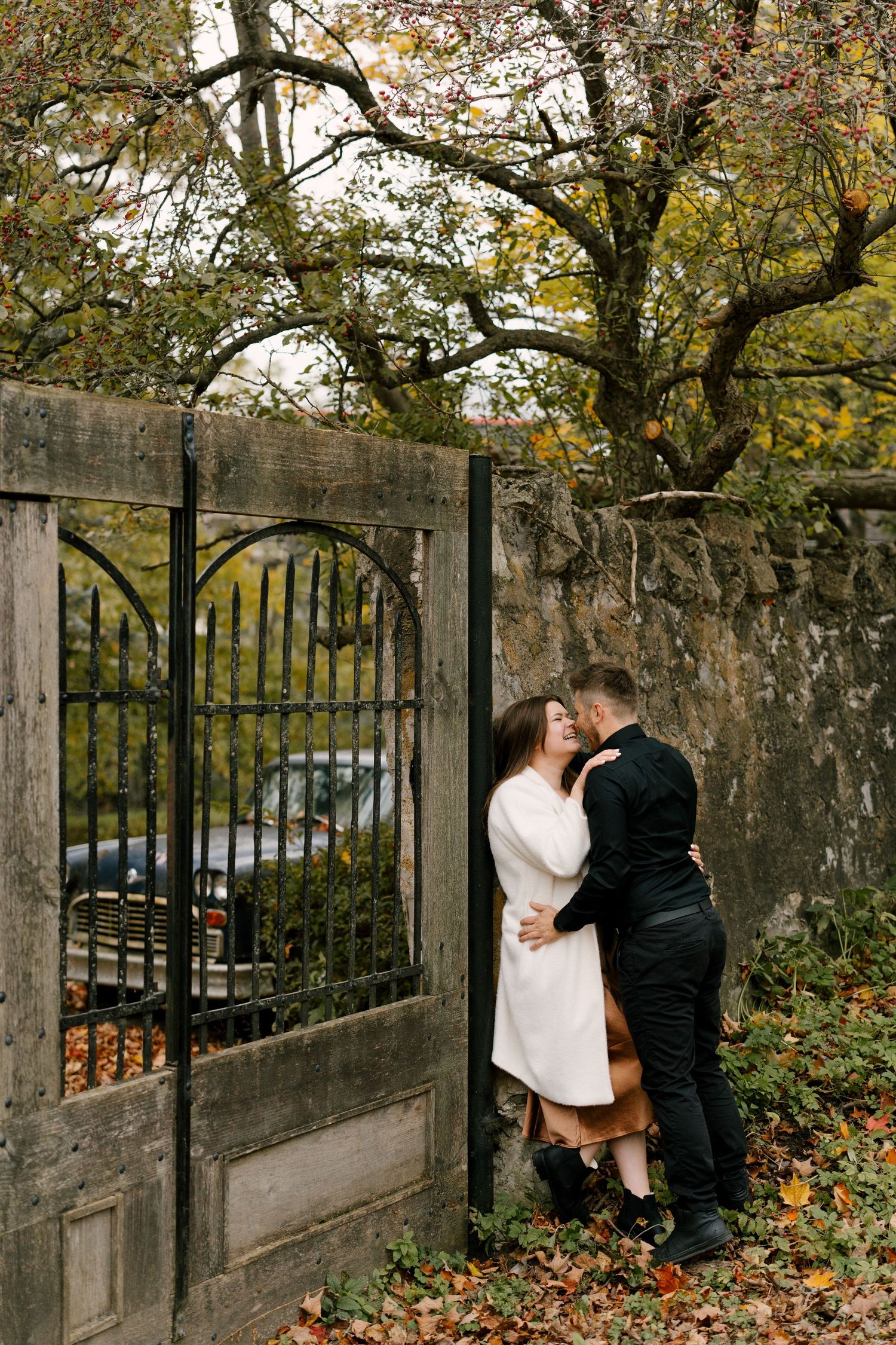 Couple embracing and kissing beside an antique wooden gate surrounded by colorful autumn leaves and trees.