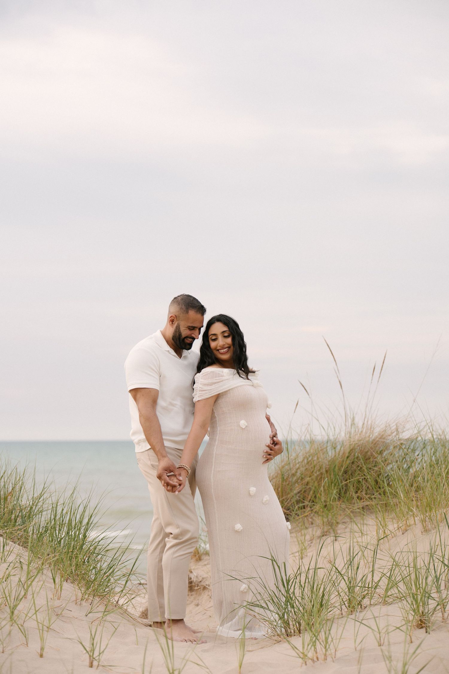 Pregnant couple in neutral tones holding hands on sandy beach dunes by the ocean.
