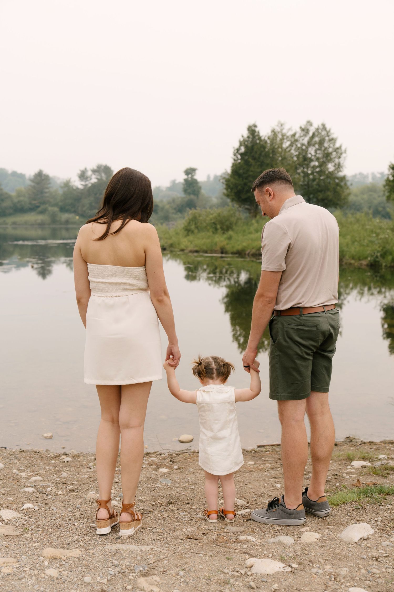 Family of three holding hands facing a calm lake surrounded by lush greenery.