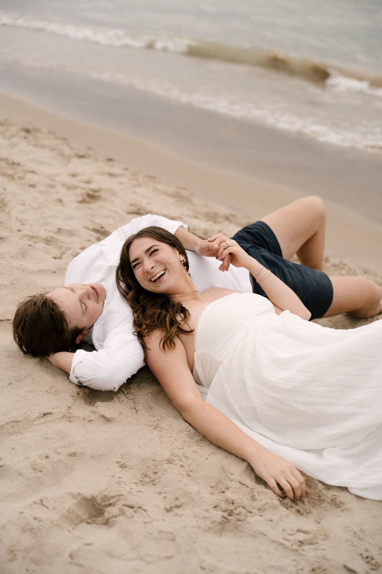 Happy couple lying on sandy beach laughing together, woman in white dress, man in white shirt and dark shorts.