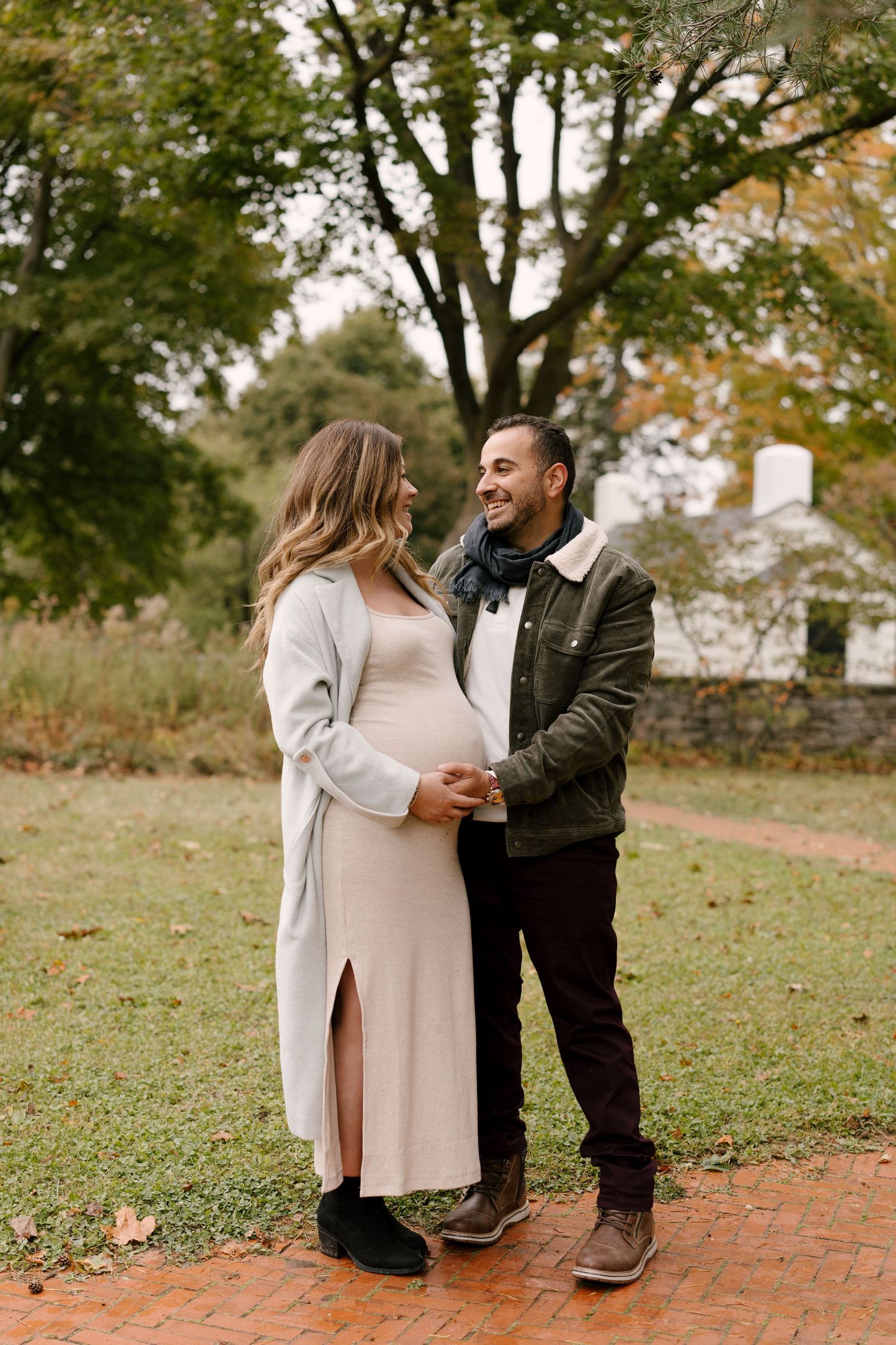 Smiling couple sharing tender moment outdoors, pregnant woman in beige dress with gray coat in fall park.