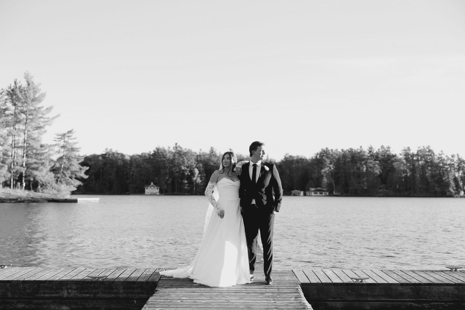Black and white photo of bride and groom standing together on a wooden dock overlooking a calm lake.