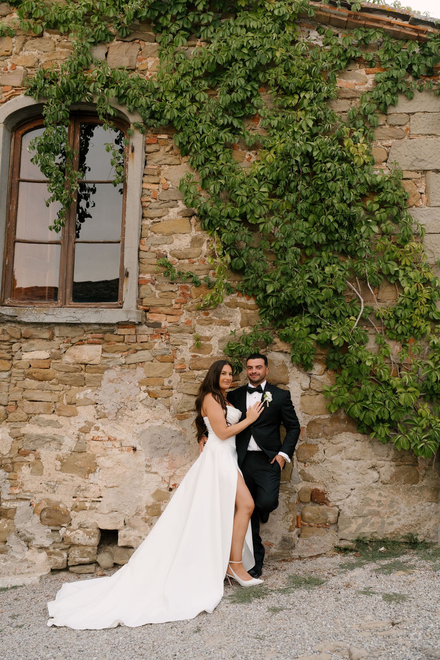 Elegant bride and groom pose against rustic stone wall covered in ivy at Italian villa.