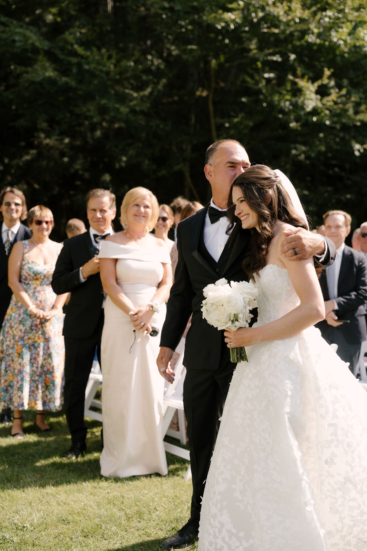 Bride in white ball gown walks down outdoor aisle with father in tuxedo, guests watching ceremony on sunny day.