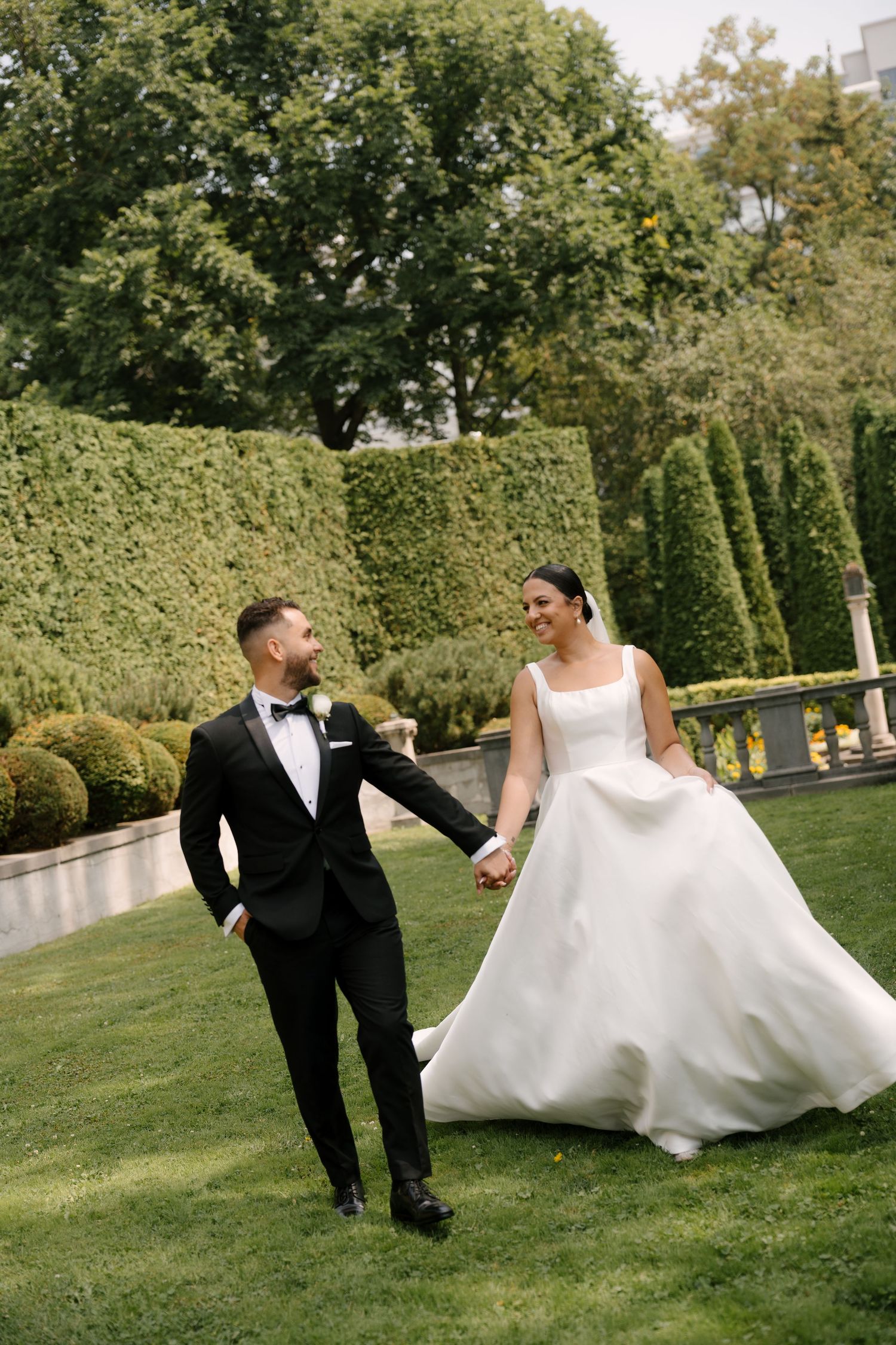 Smiling bride in a white ball gown and groom in black tuxedo walking hand-in-hand across a lush green lawn at Parkwood Estates.