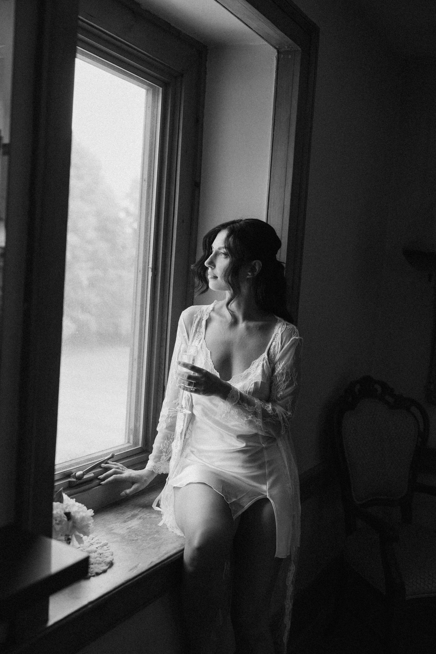 Black and white photo of bride in lace robe sitting on windowsill gazing outside thoughtfully at Roseville Estates wedding venue.