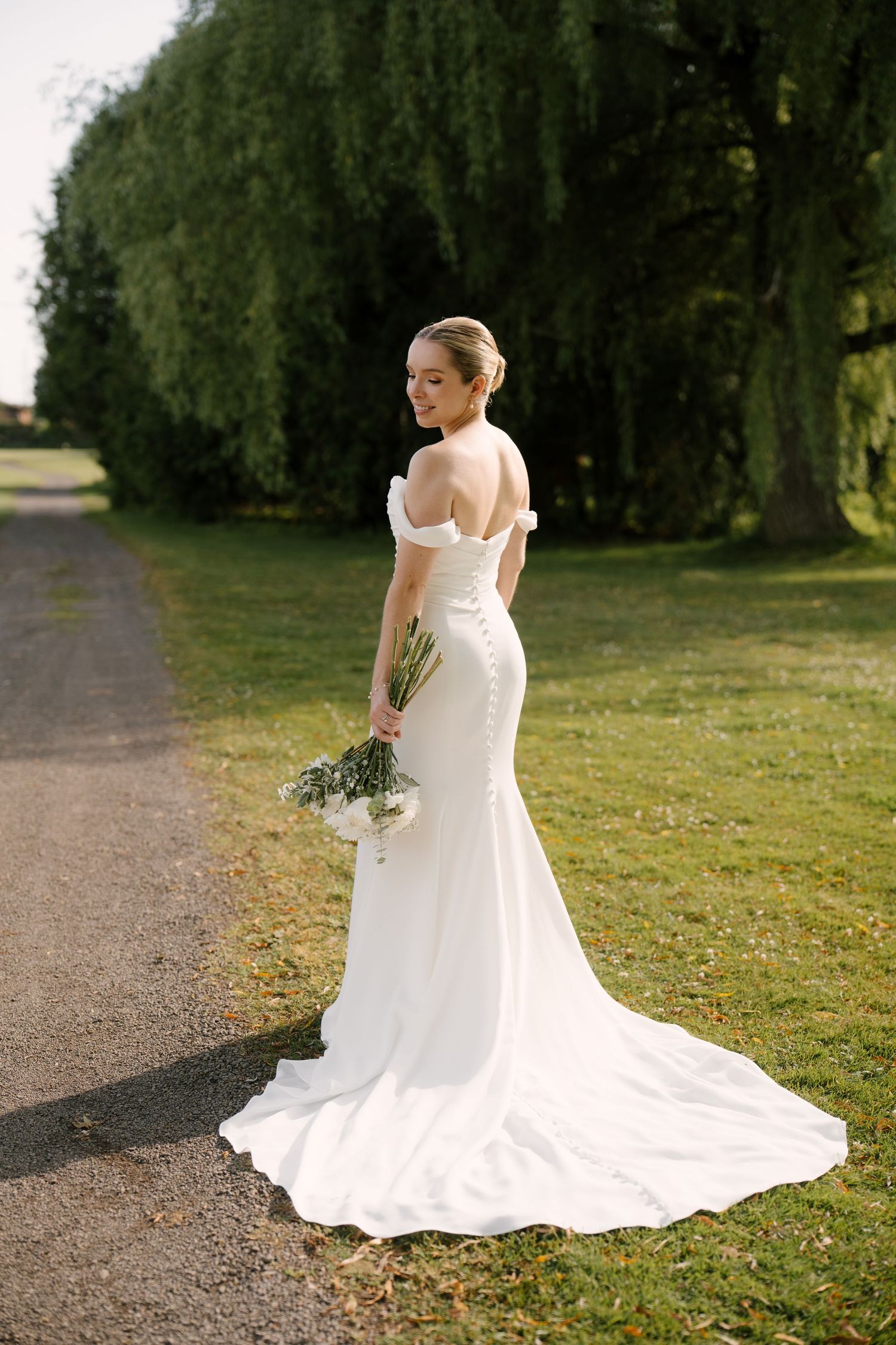 Bride in fitted off-shoulder wedding dress with button back train poses on garden pathway at Guelph golf club.