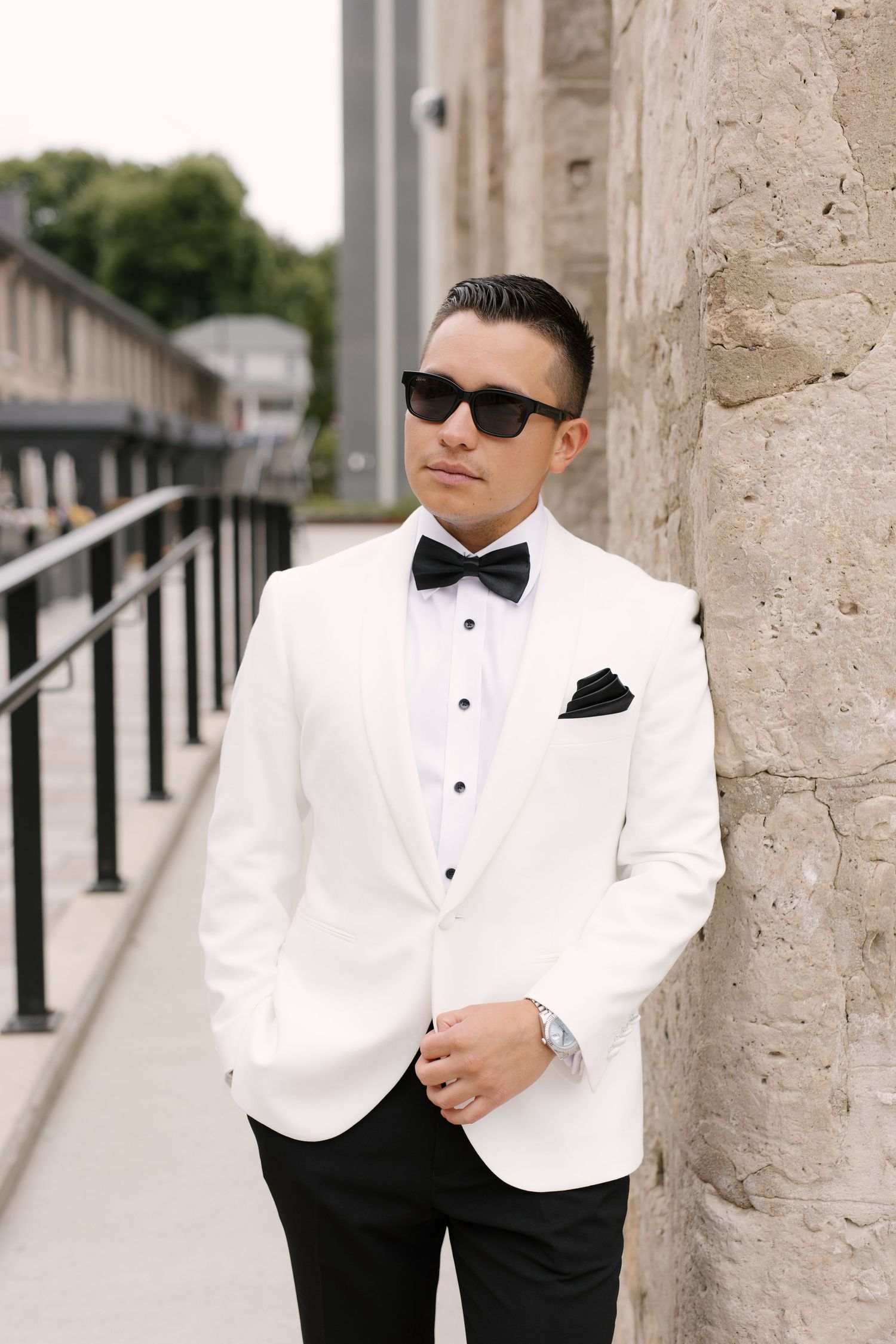 Stylish groom in white tuxedo jacket and sunglasses leans against a stone wall outside Tapestry Hall.