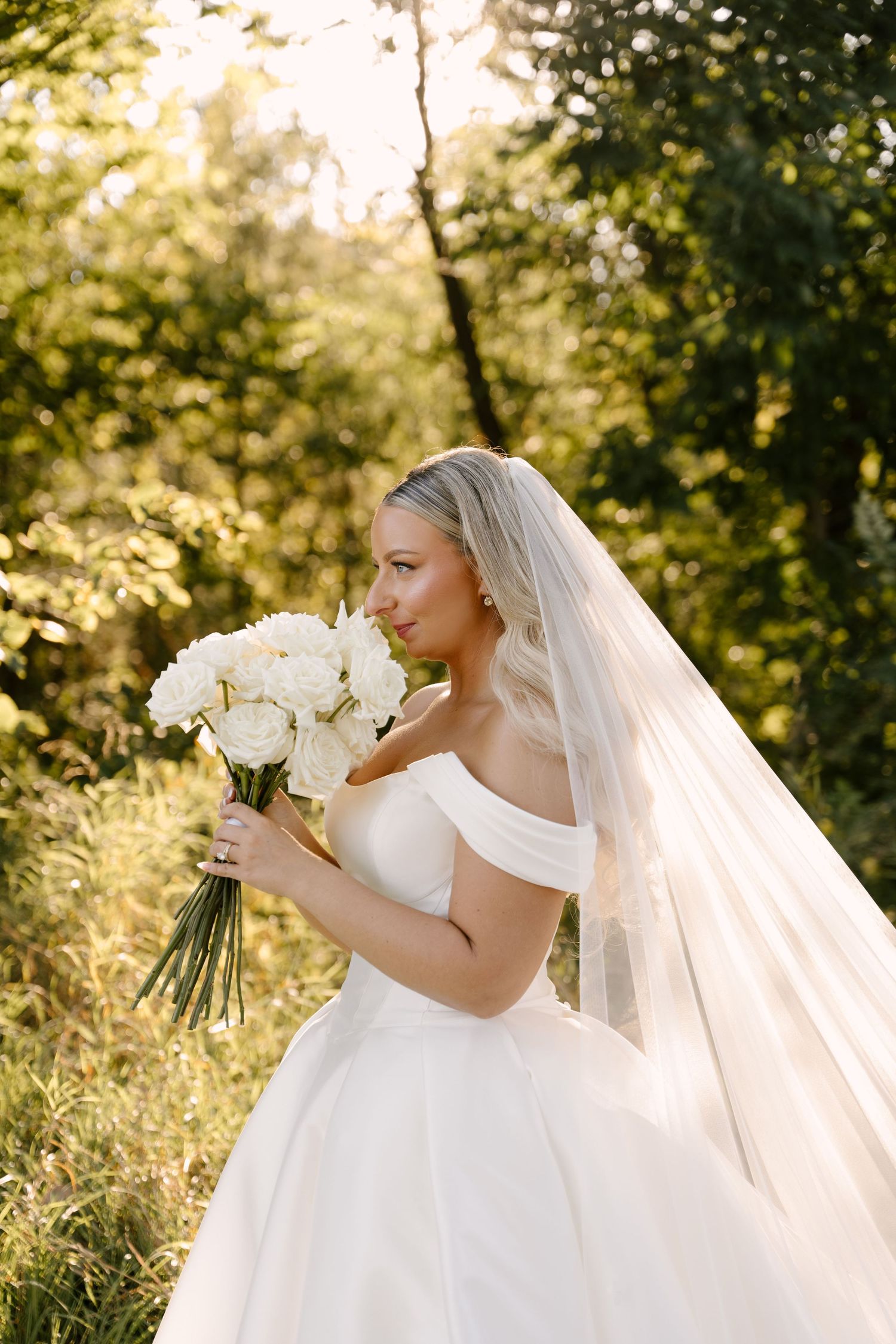 Elegant bride in off-shoulder gown holds white peony bouquet in sunlit garden setting at Whistle Bear wedding venue.