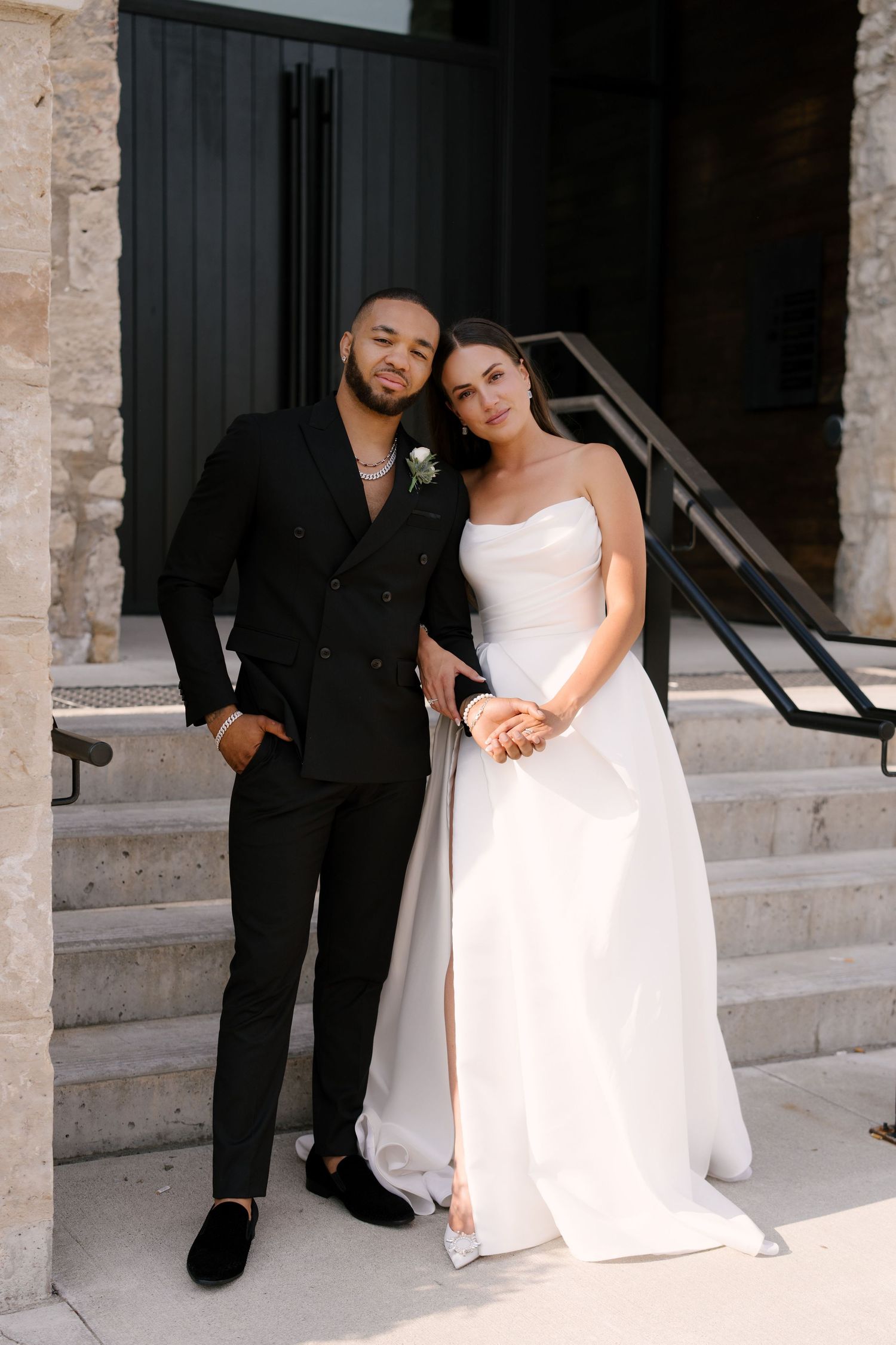 Bride in strapless white gown leans on groom in black double-breasted suit on stone building steps outside Tapestry Hall wedding venue.