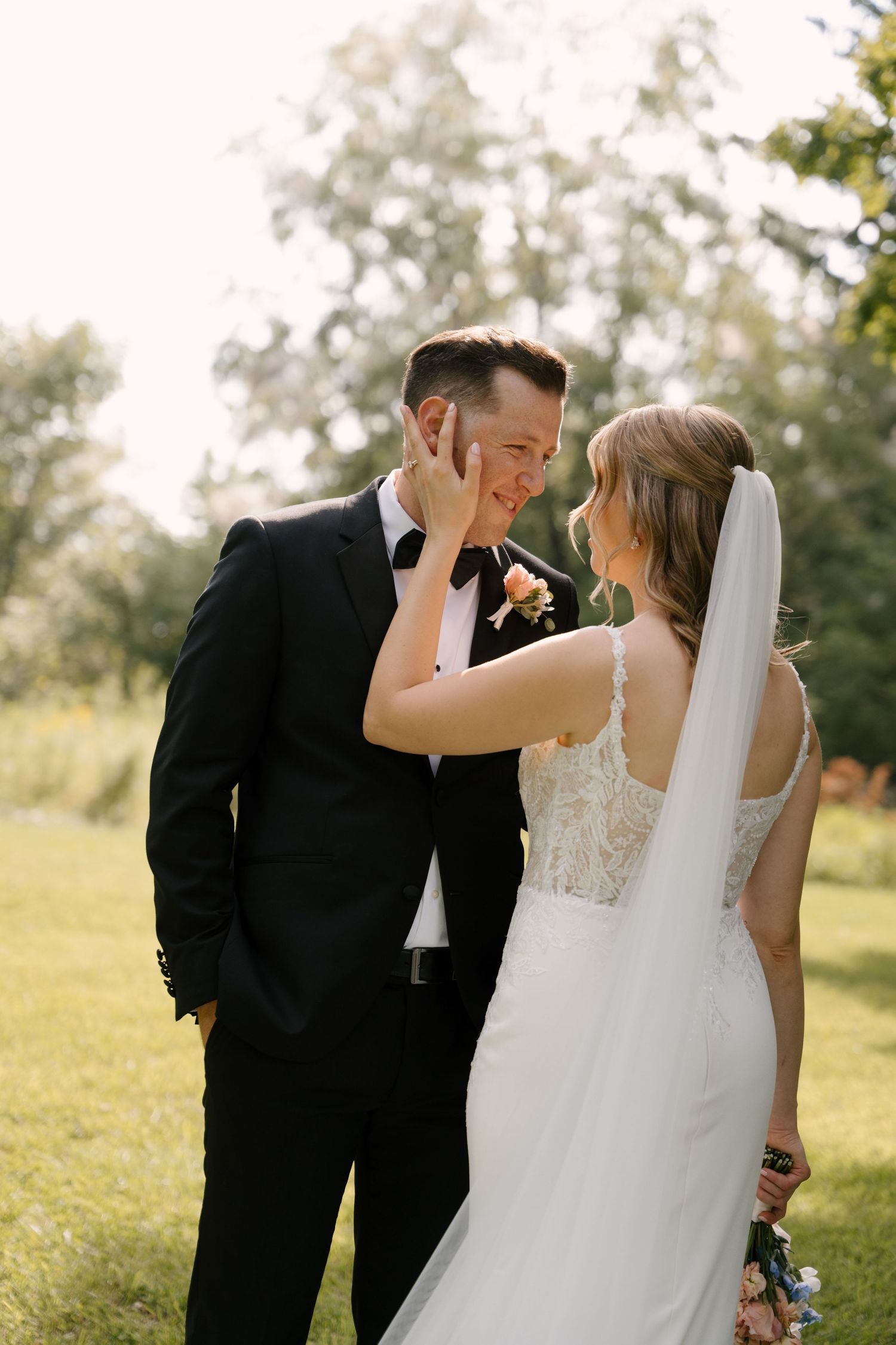 Bride tenderly touches grooms face as they share an intimate moment outdoors in golden sunlight on their wedding day.