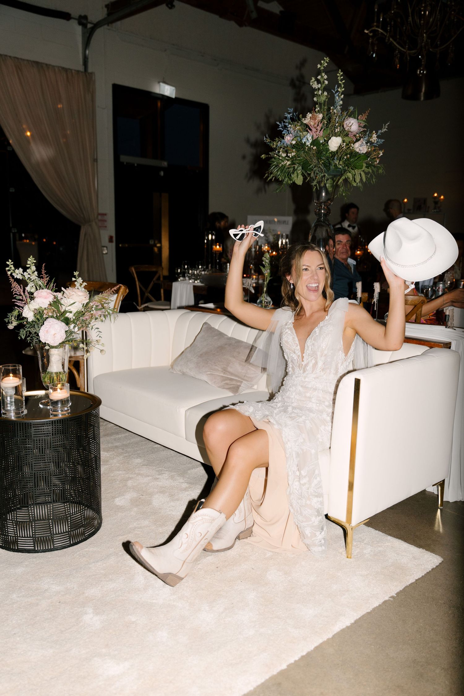 Bride in lace dress and cowboy boots celebrates on white sofa, holding hat and sunglasses at St Marys Golf Club wedding reception.