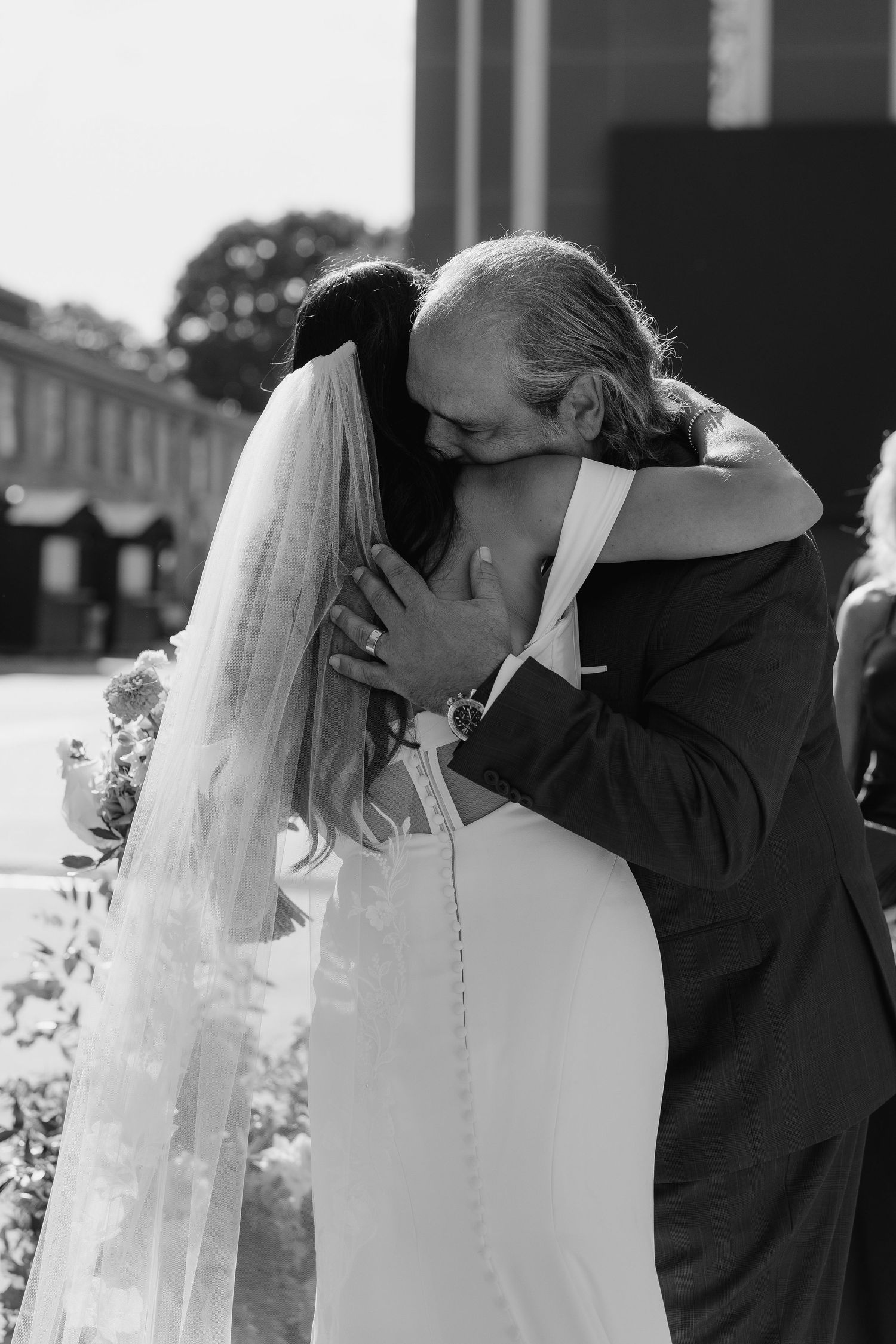 Emotional black and white photo of a bride in a veil sharing a heartfelt hug with father at her wedding ceremony.