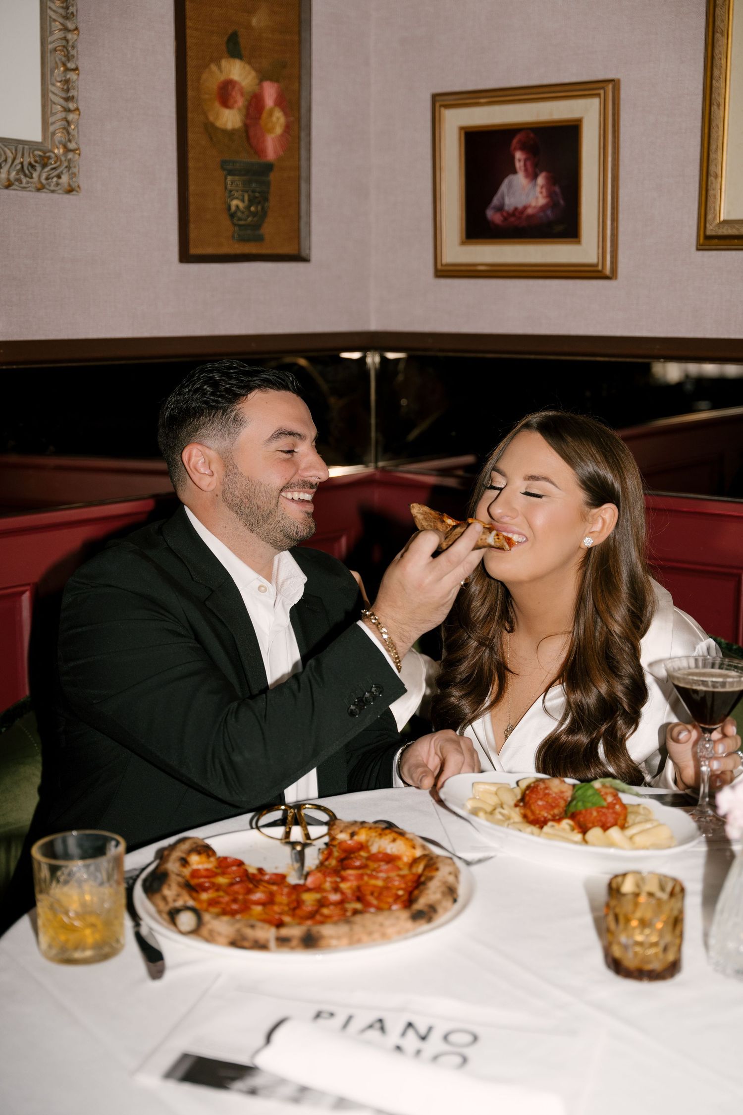 Joyful couple sharing pizza at Piano restaurant, man feeding woman as they laugh at candlelit table.