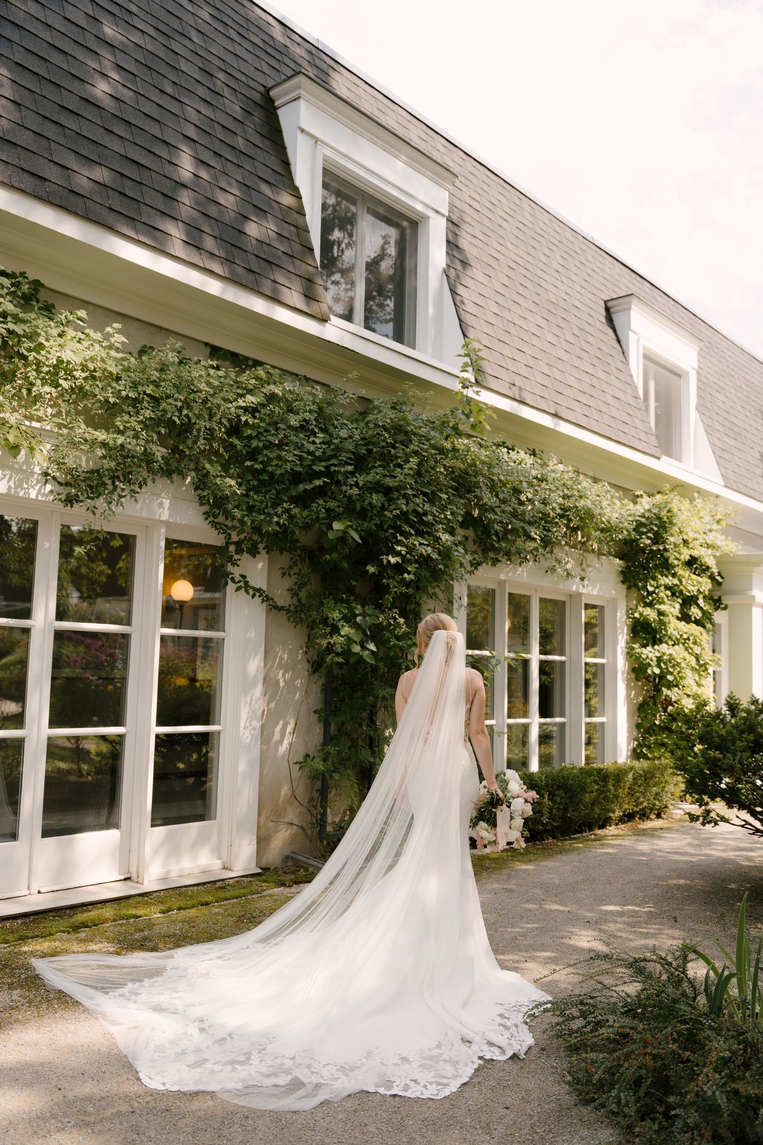 Bride in white gown with long lace-edged cathedral veil stands before ivy-covered venue exterior at Langdon Hall.