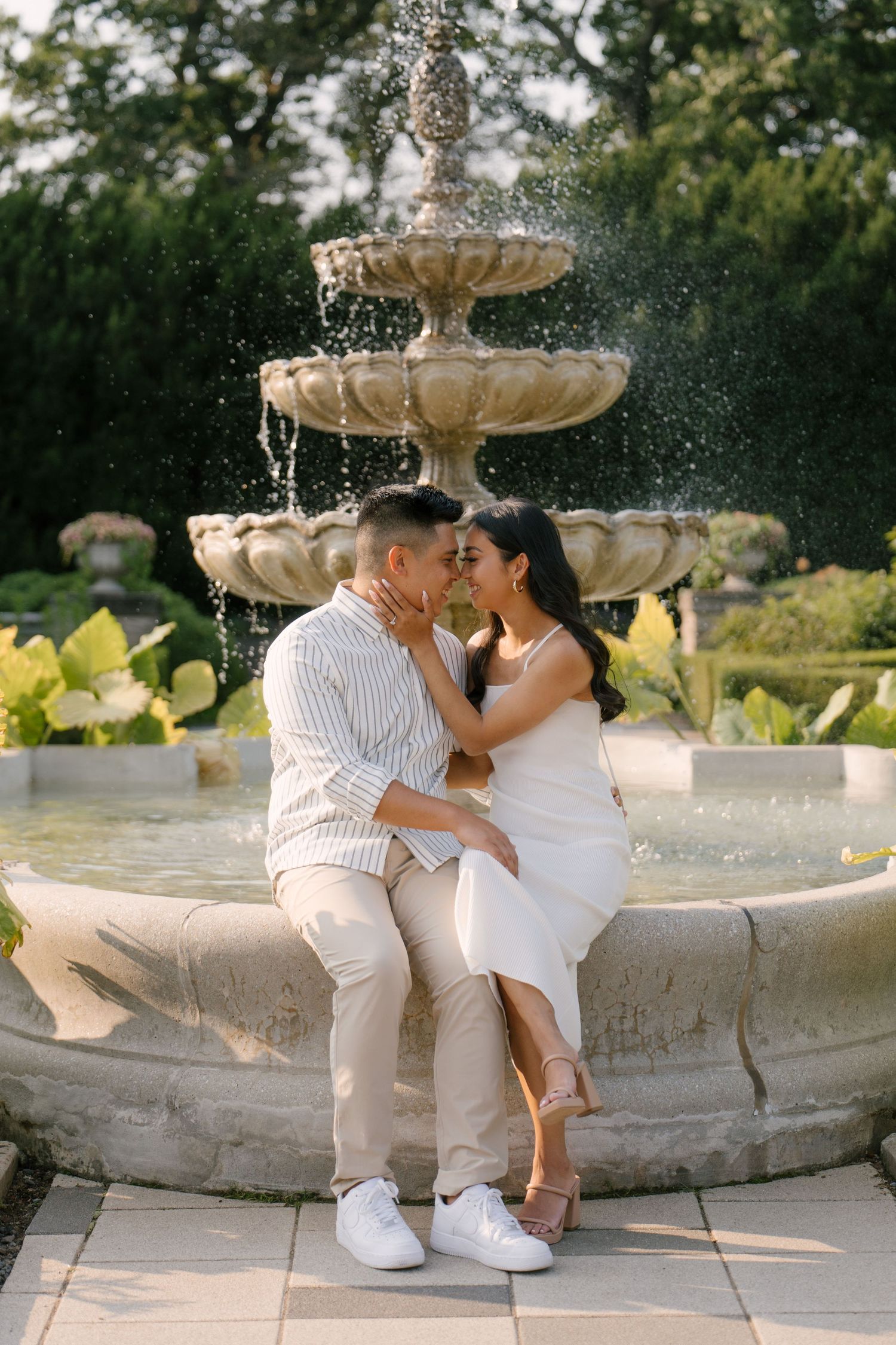 Couple sharing a tender moment seated beside an elegant stone tiered fountain in a lush garden.