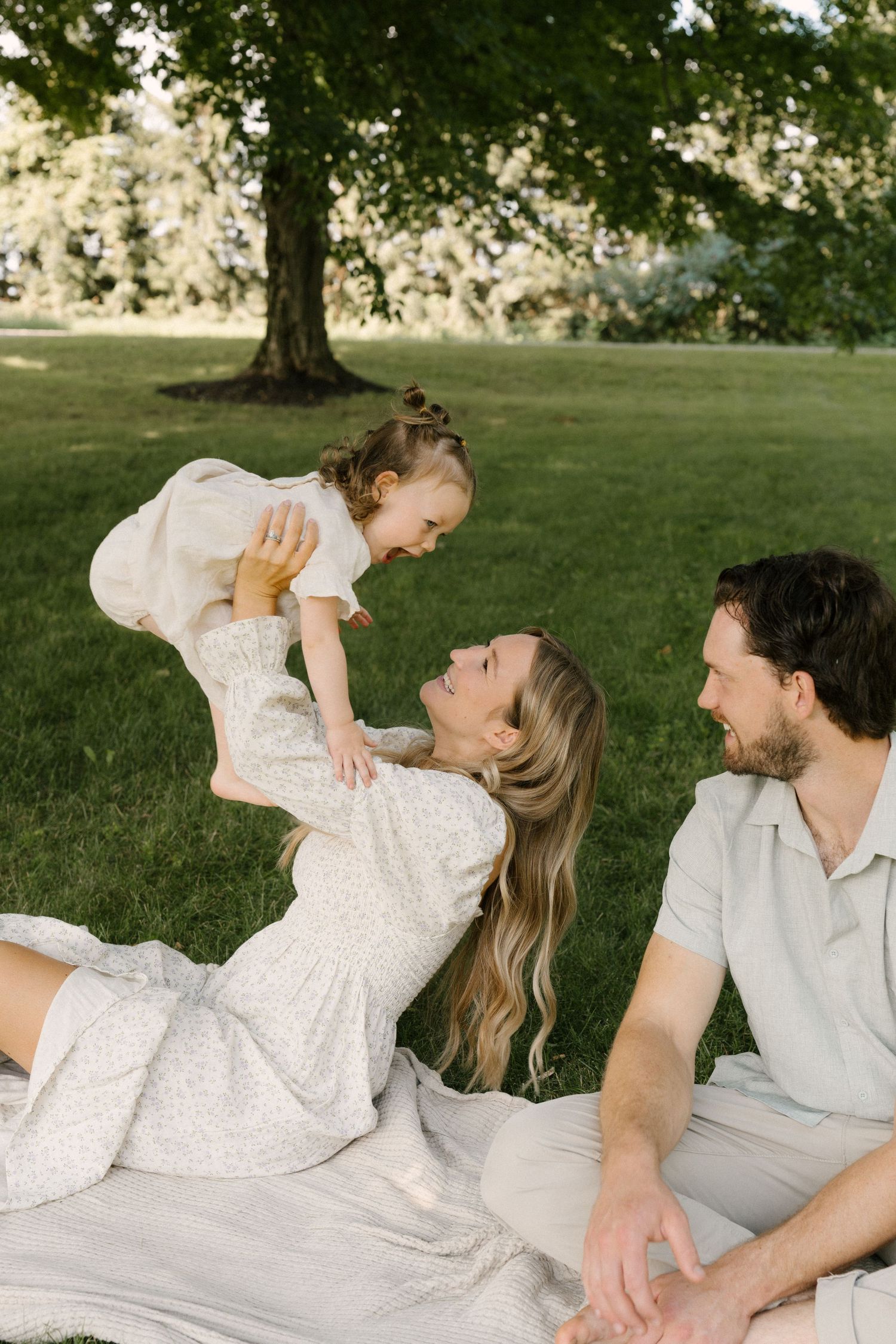 A laughing mother lifts her giggling toddler while lying on a blanket as her husband smiles nearby.