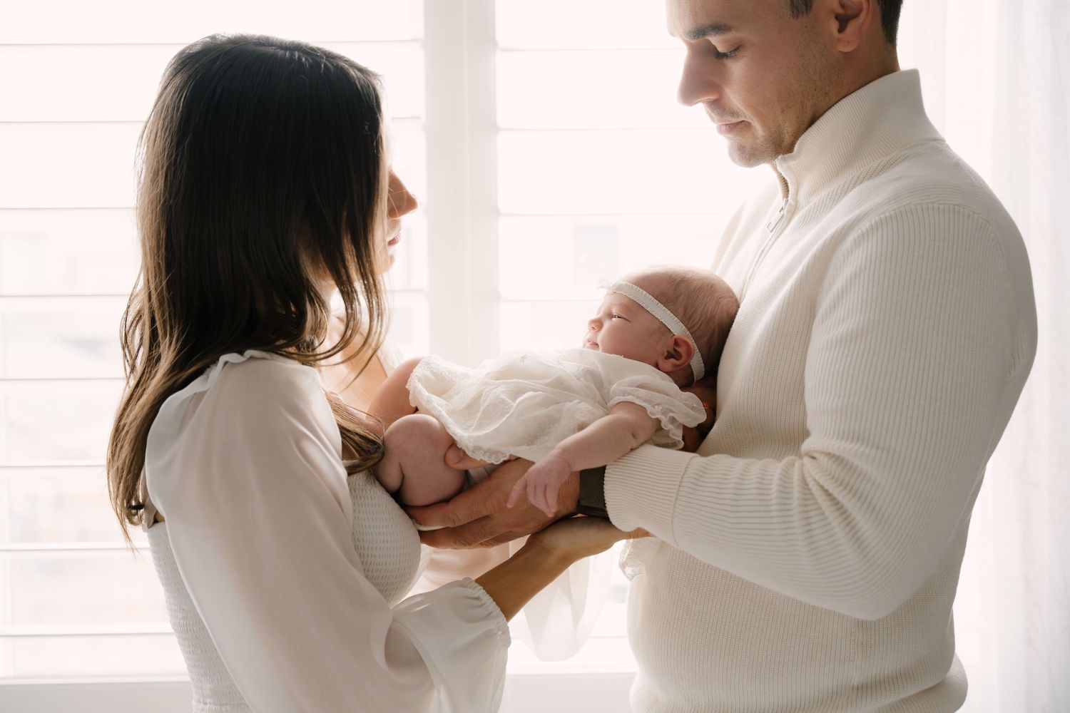 Parents in white attire gently cradling their sleeping newborn by a bright window.