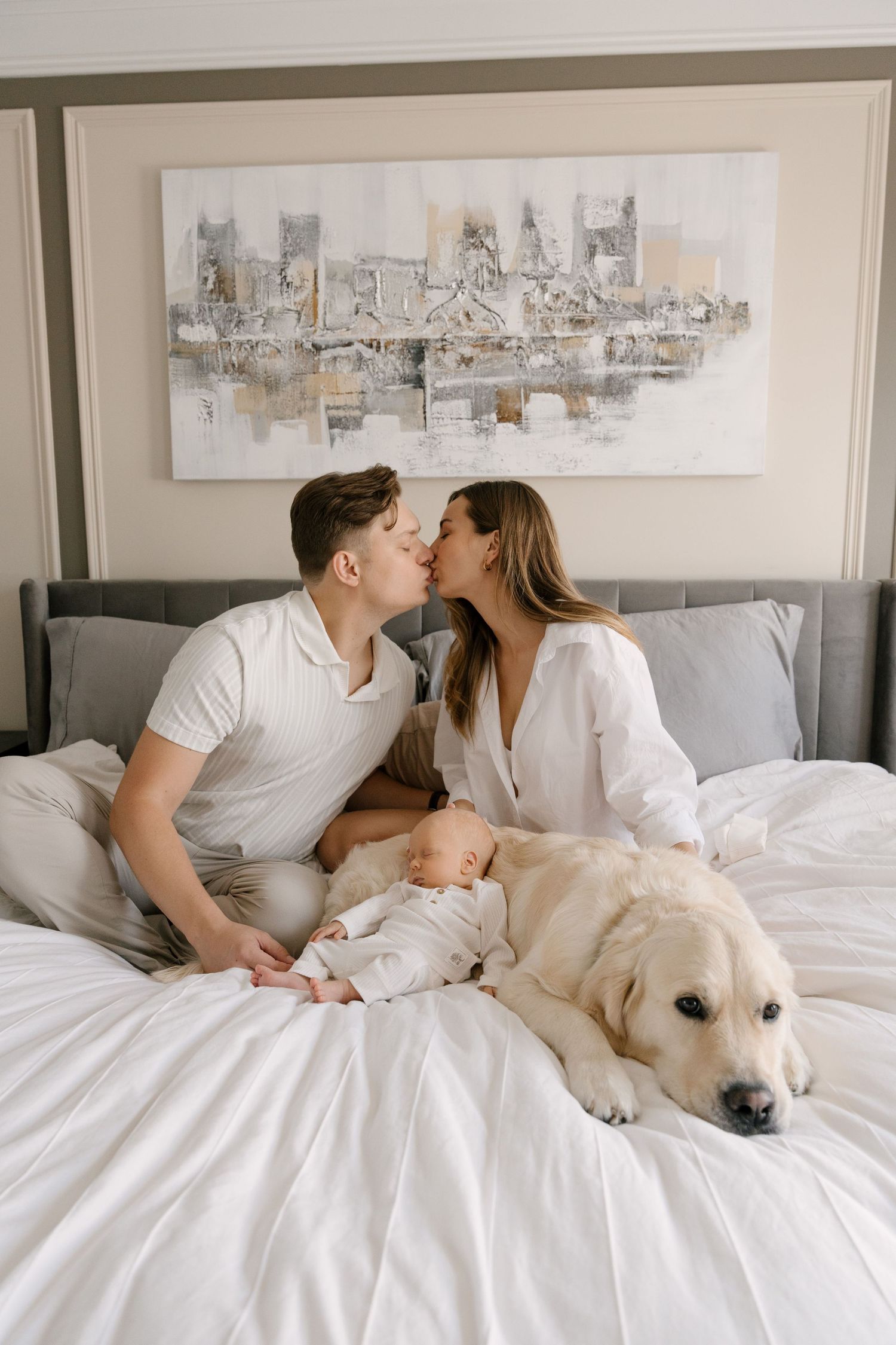 A couple kisses on a white bed with their newborn baby and golden retriever in a cozy bedroom.