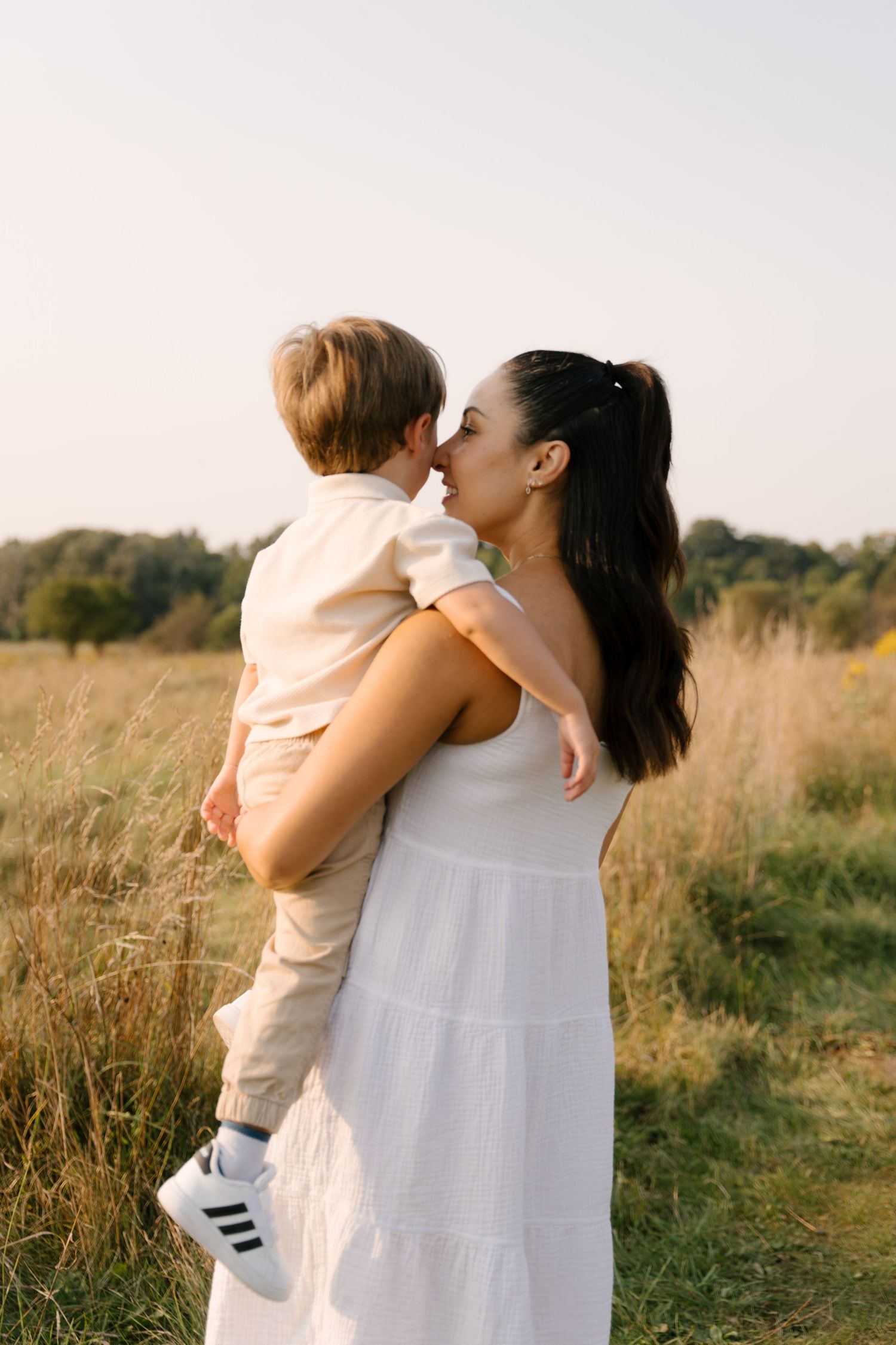 Dark-haired mother in white dress tenderly kisses her young son while holding him in a golden field.