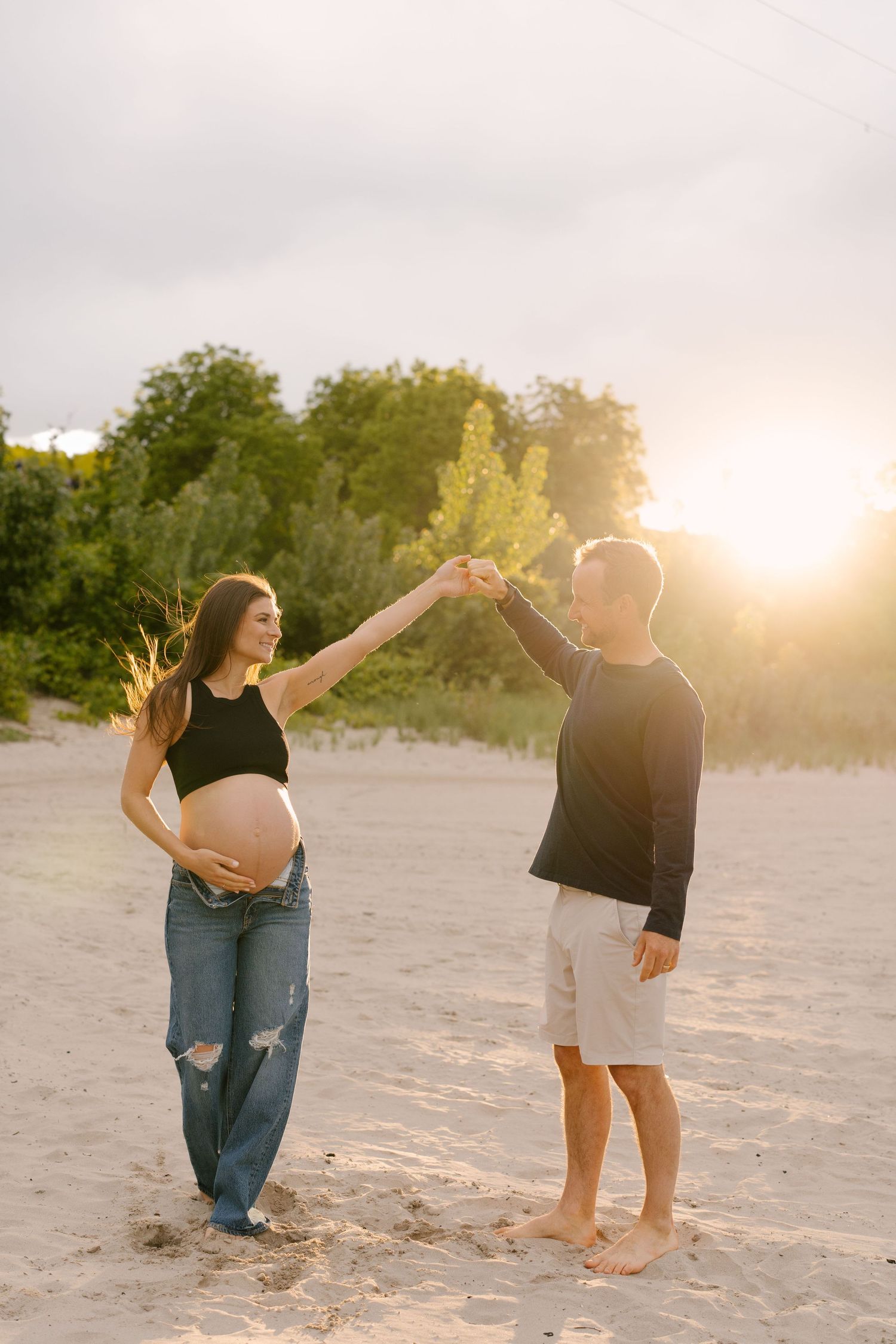 Pregnant woman in black crop top holds partners raised hand on sunlit sandy beach during golden hour.