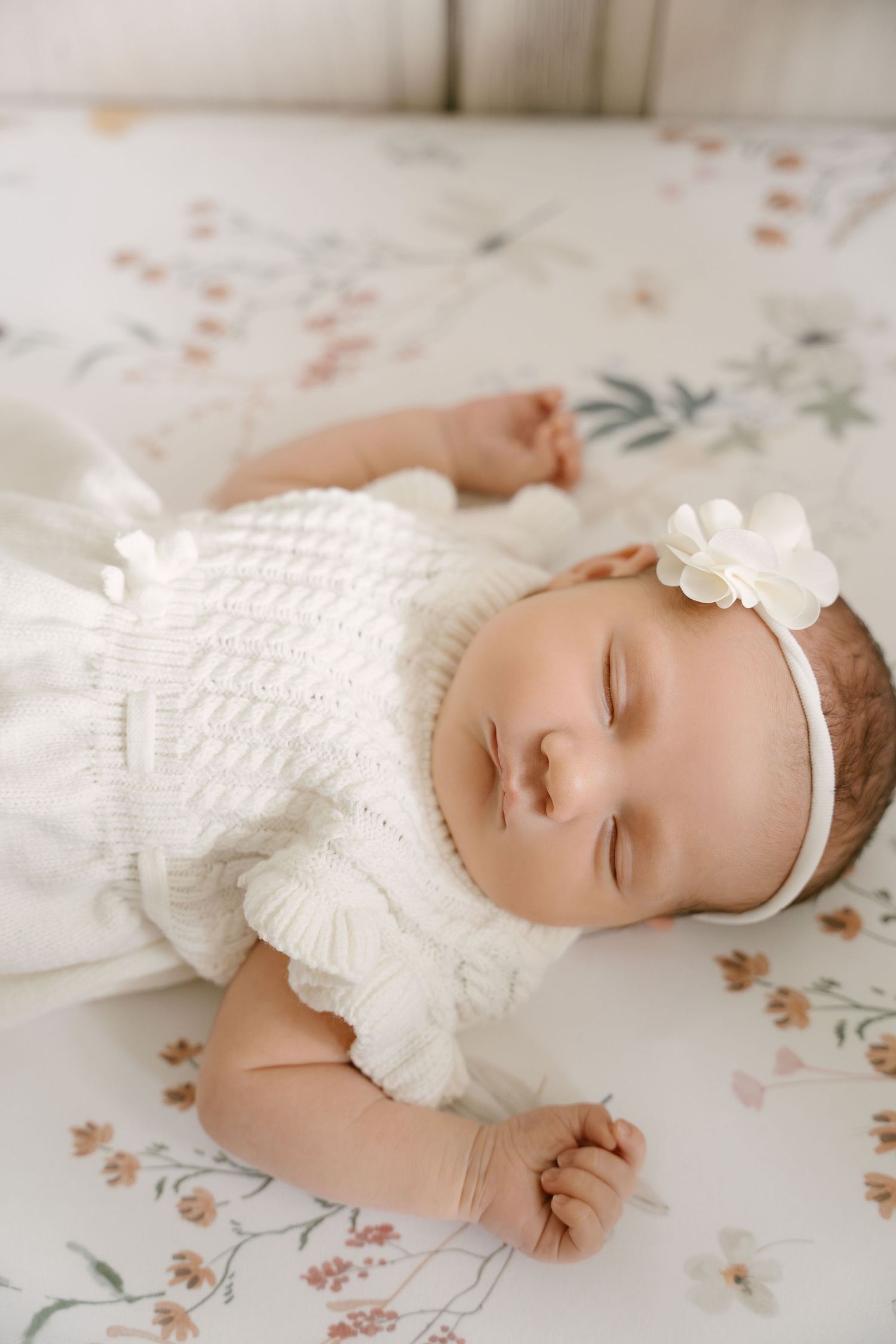 Close-up of sleeping newborn baby girl in white knit romper with flower headband on floral bedding.