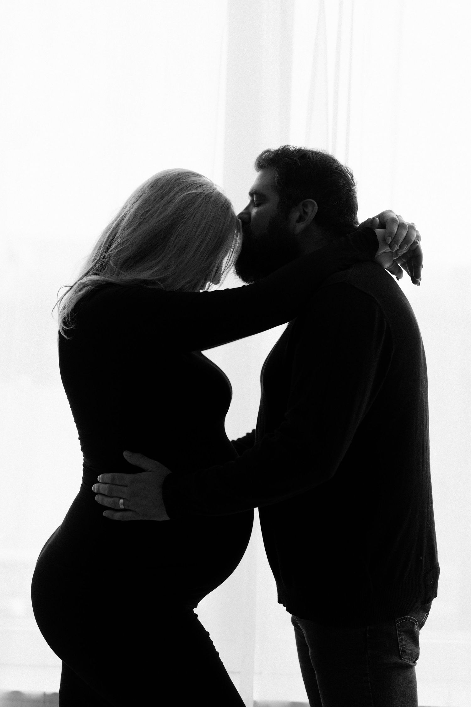 Black and white silhouette of pregnant couple kissing tenderly in front of bright window.