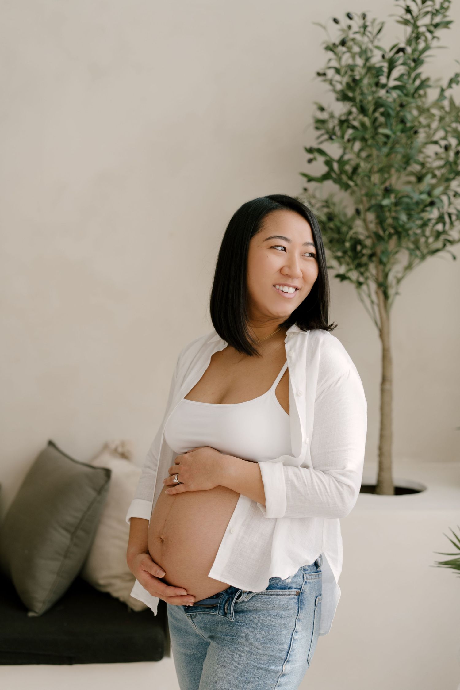Smiling pregnant Asian woman in white top and jeans cradles her baby bump in a bright modern room.