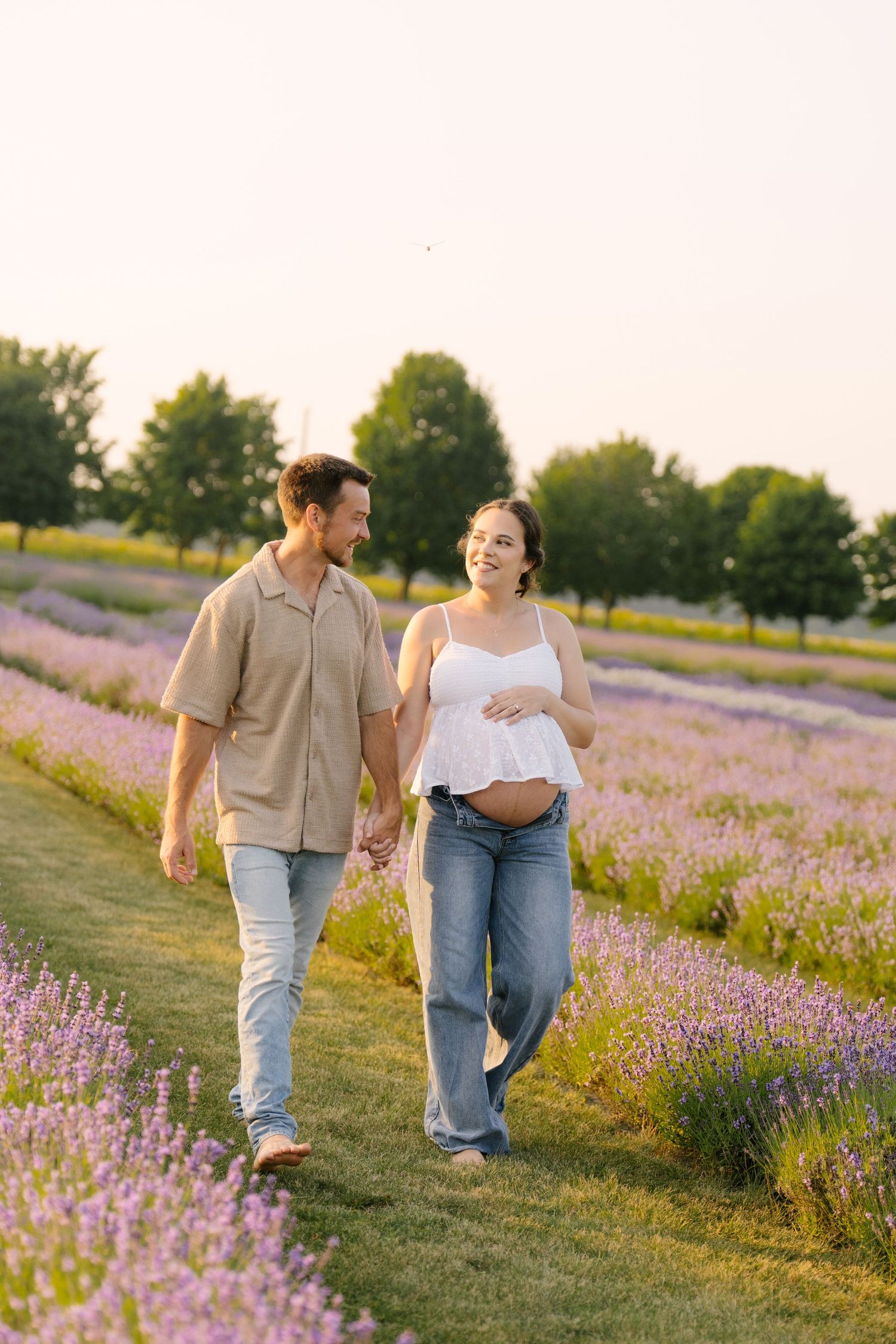 Laughing pregnant woman holding baby bump while walking hand-in-hand with partner through lavender field.