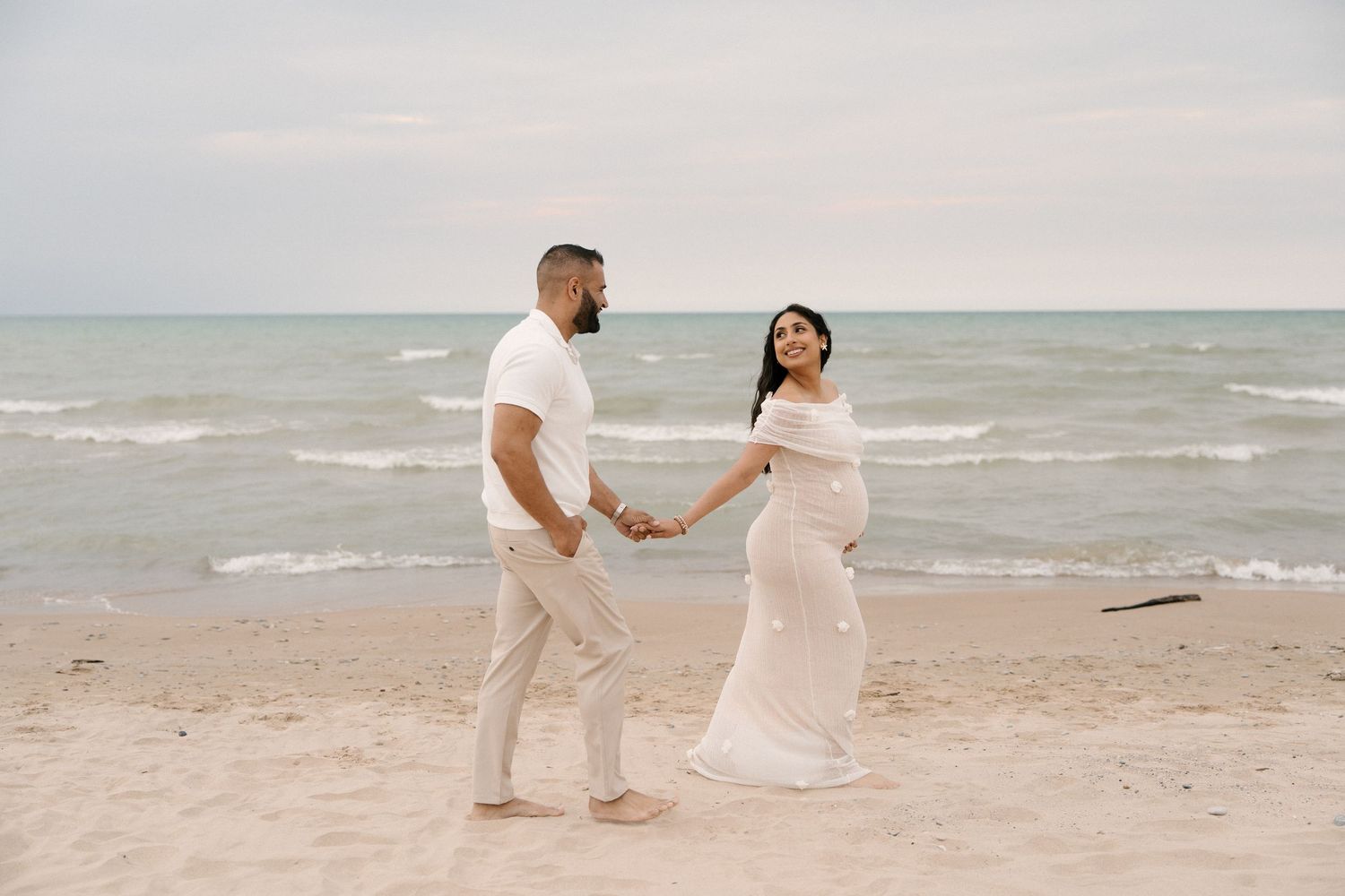 Expecting couple holding hands on sandy beach, woman in white maternity gown, man in white outfit.