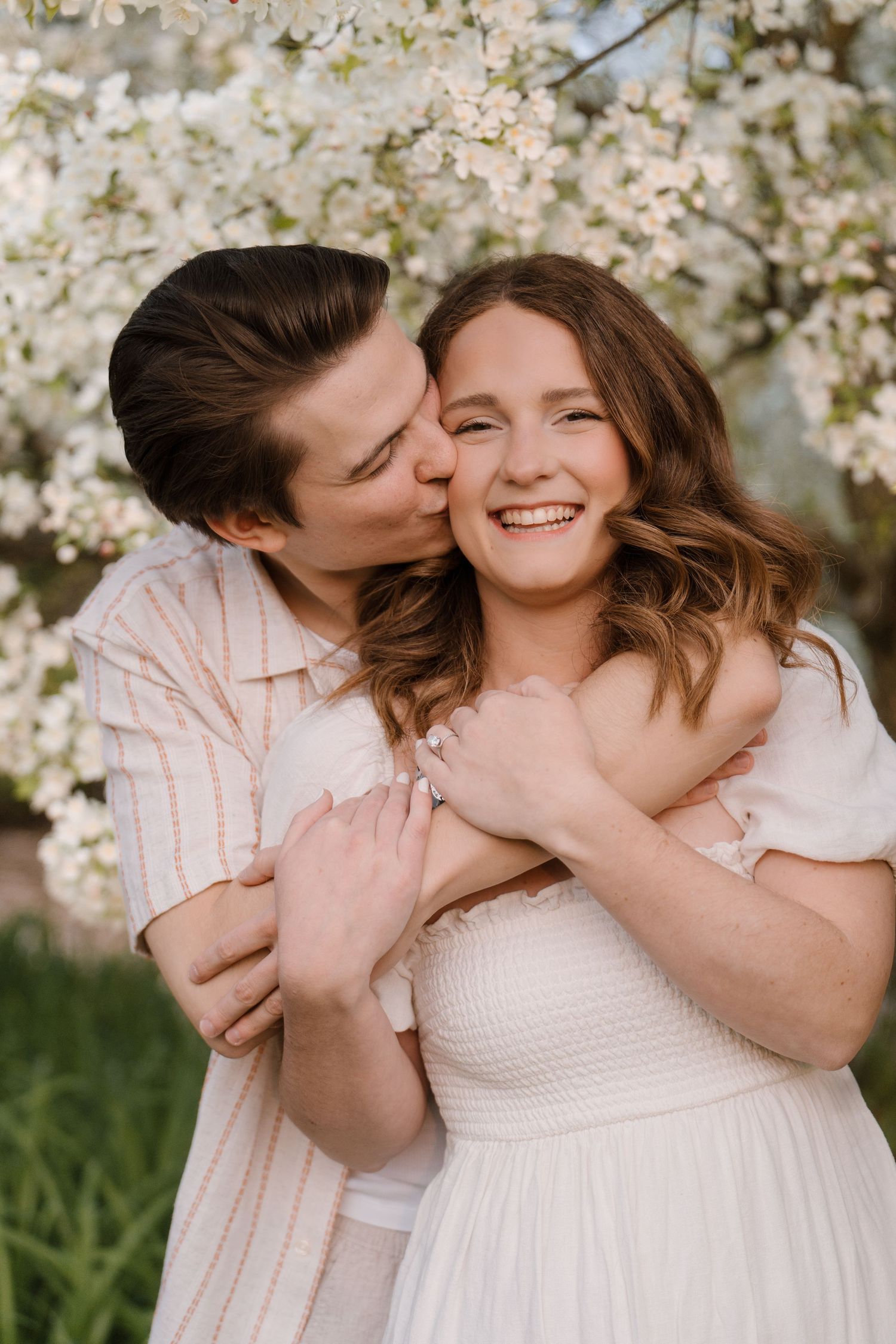 Man kissing woman on cheek while hugging her from behind as she laughs joyfully near white blossoms.