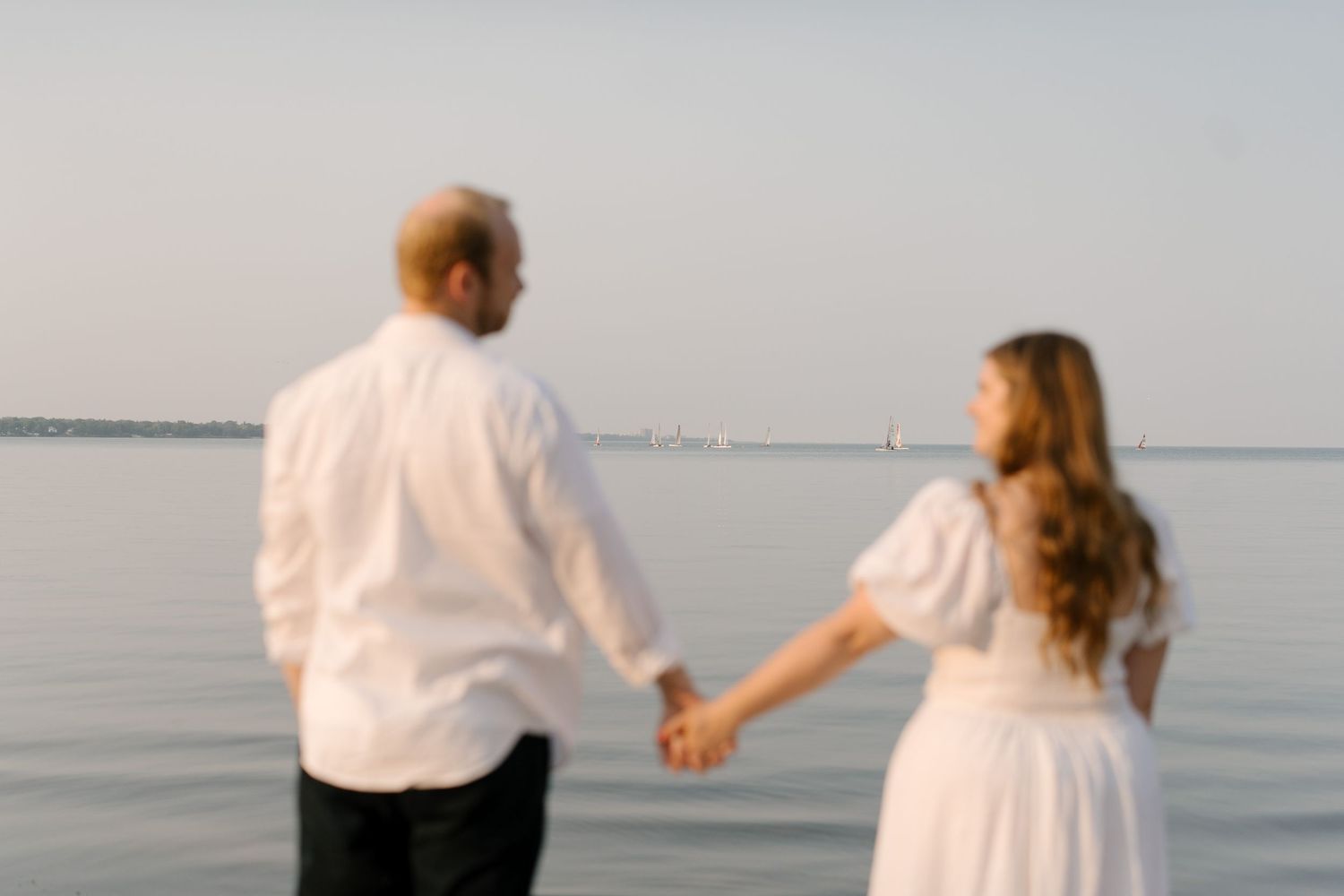 Couple holding hands facing the calm ocean at dusk, both dressed in white outfits on a sandy beach.