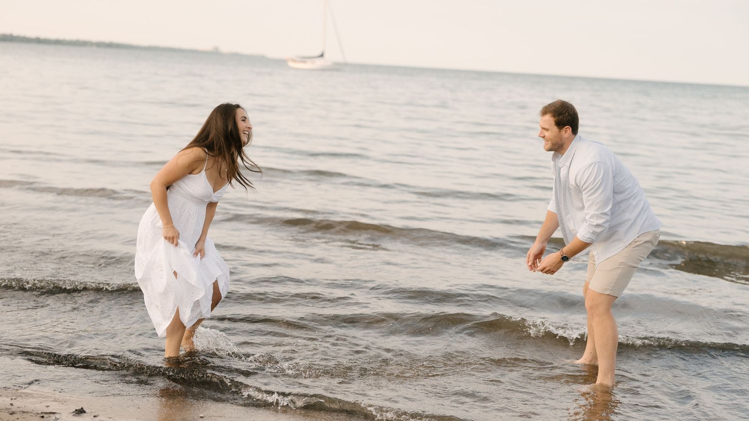 Couple playfully splashing in ocean waves at beach, woman in white dress laughing with man in white shirt.