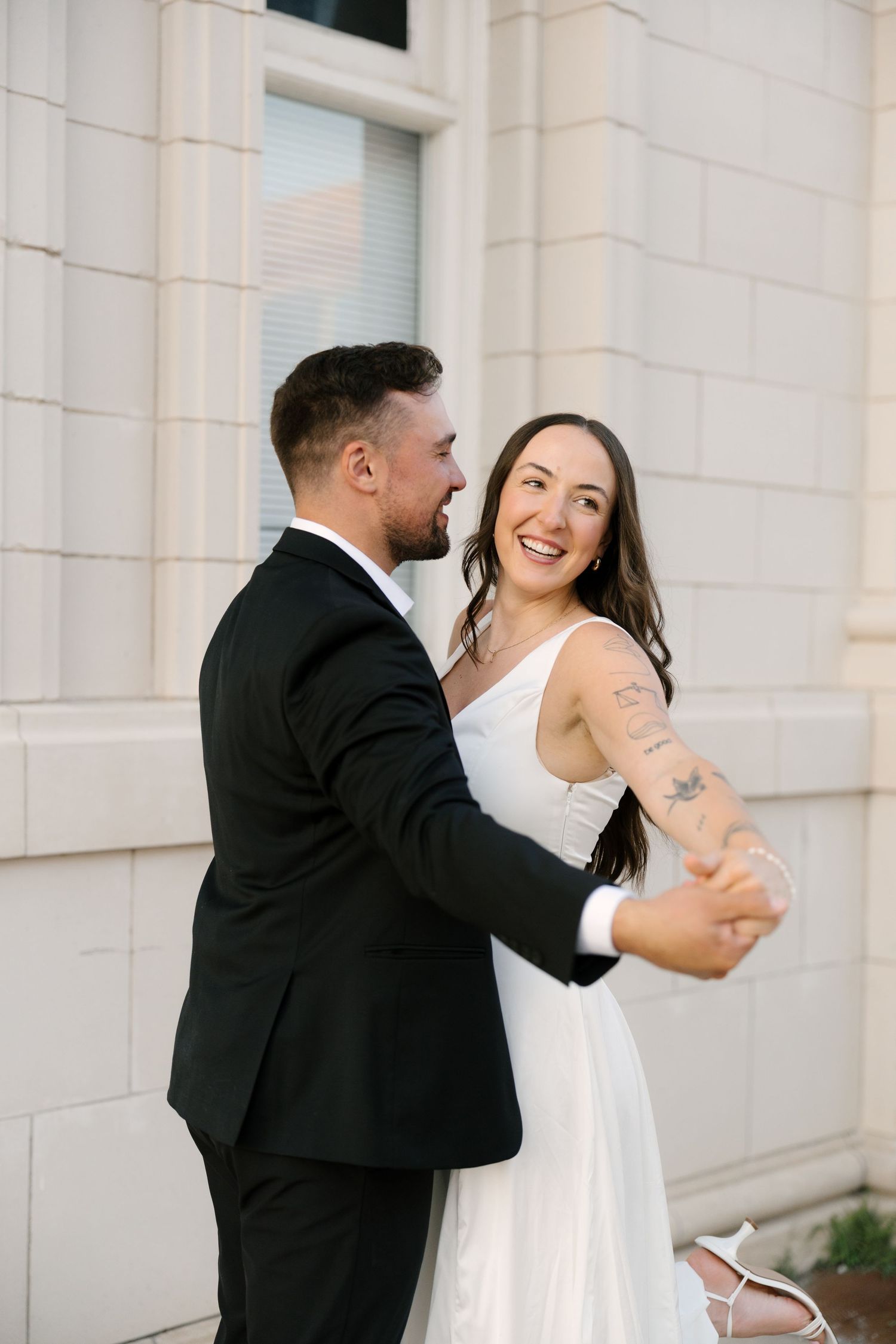Joyful bride in white dress dancing with groom in black suit outdoors near stone building.
