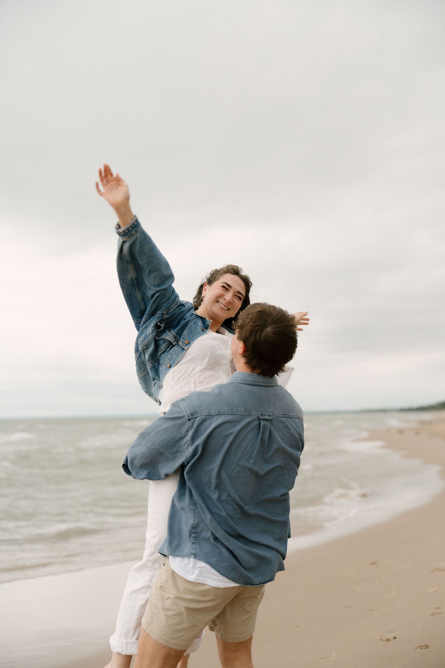 Man lifting smiling woman with arms raised into the air on a cloudy beach with ocean waves behind.