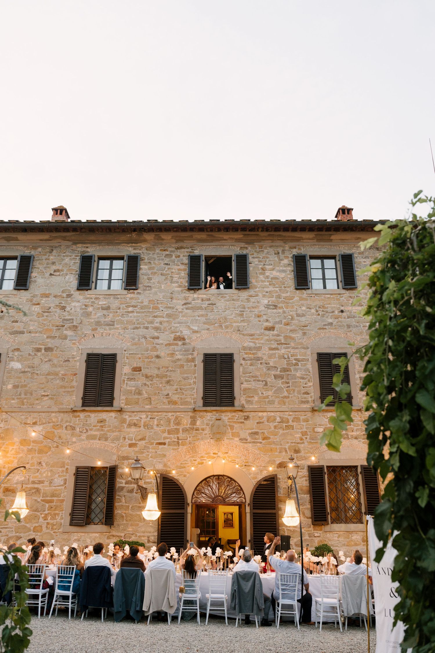 Outdoor wedding reception dinner at dusk in front of historic stone Tuscan villa.