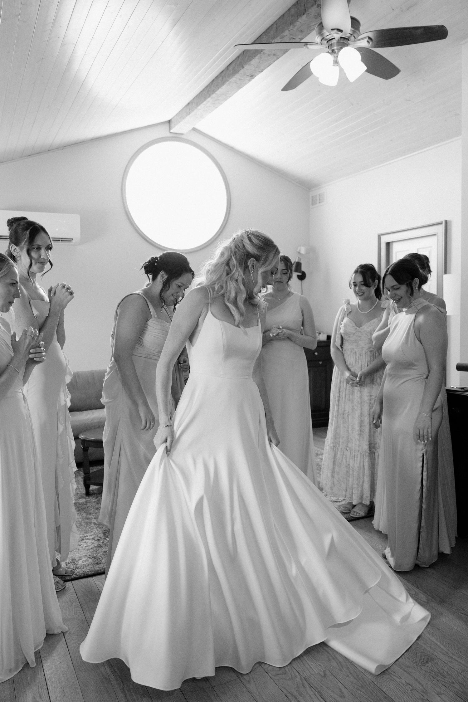 Bride in white gown twirling while surrounded by admiring bridesmaids in elegant black and white photo.