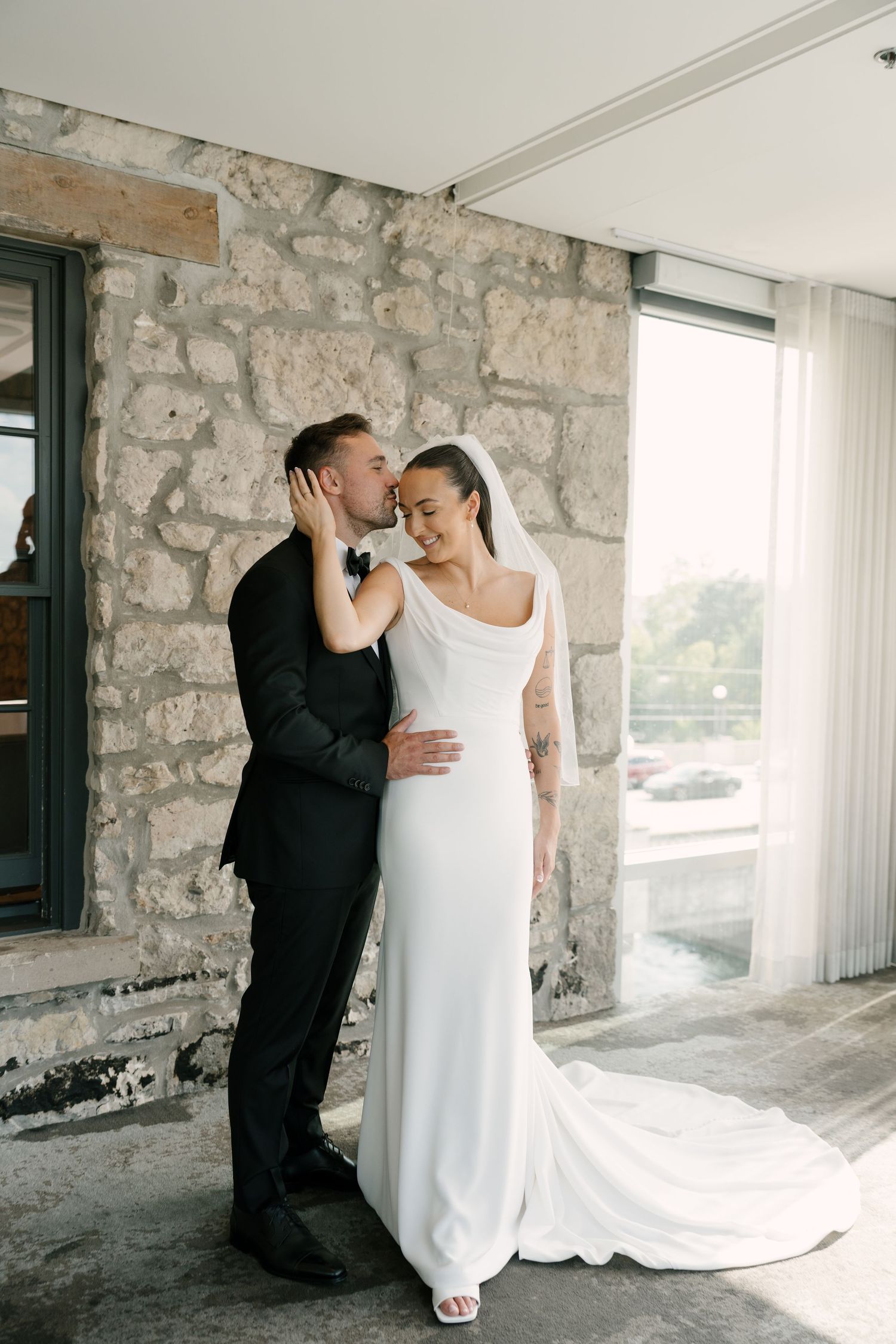 Groom in black suit kisses bride in white gown and veil against stone wall backdrop at Cambridge Mill.