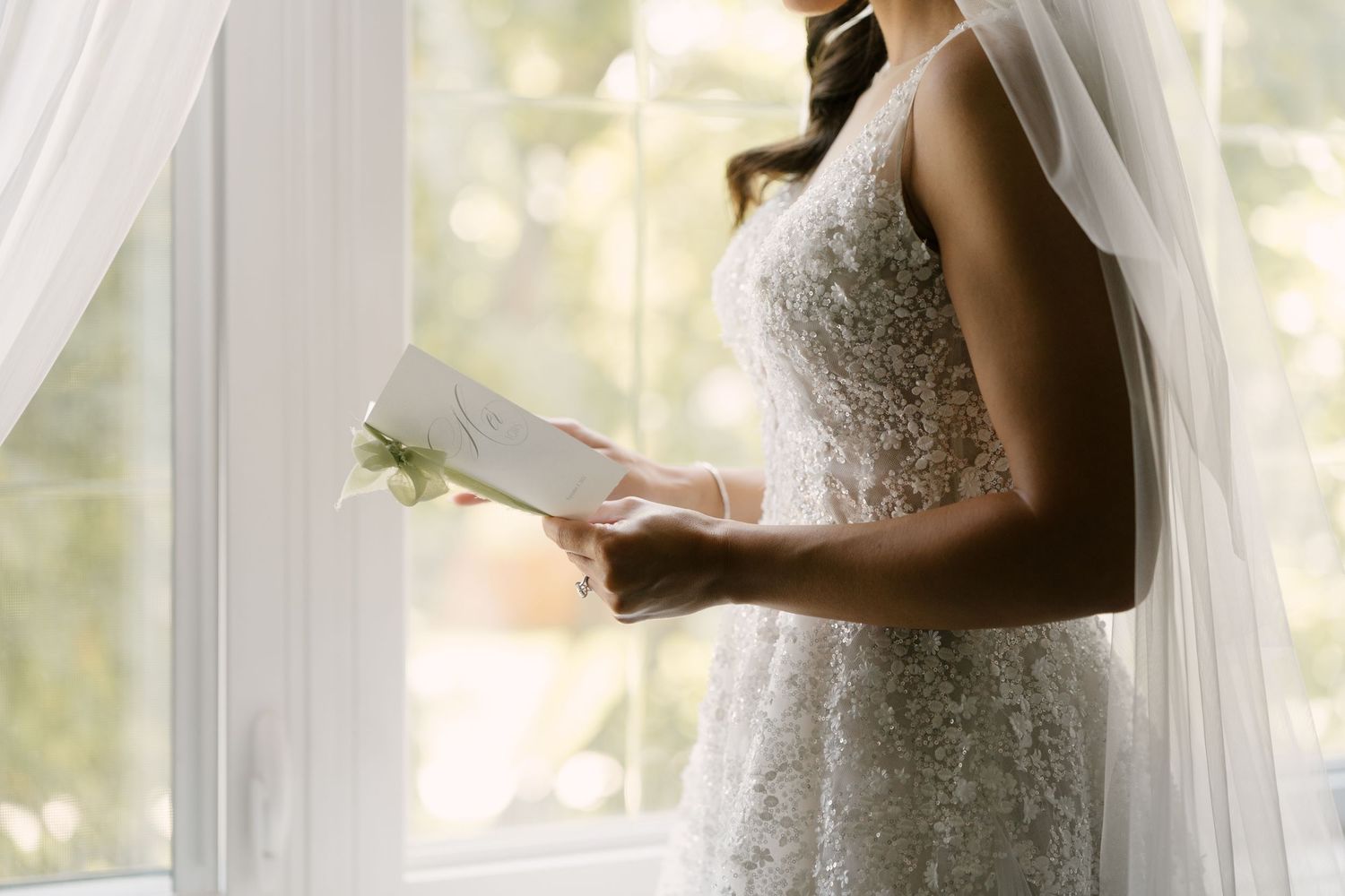 Bride in lace wedding gown and veil reading a letter by a sunlit window on her wedding day.