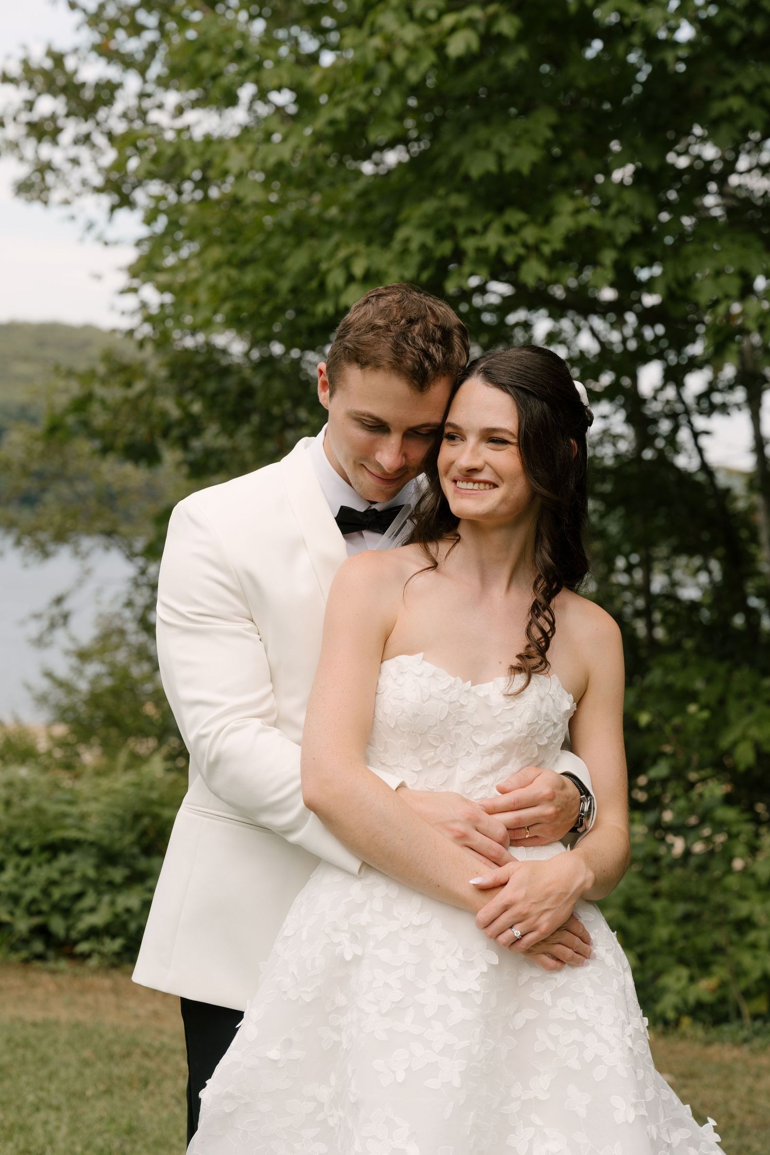 A smiling bride in a strapless lace gown embraced by a groom in a white tuxedo jacket outdoors.
