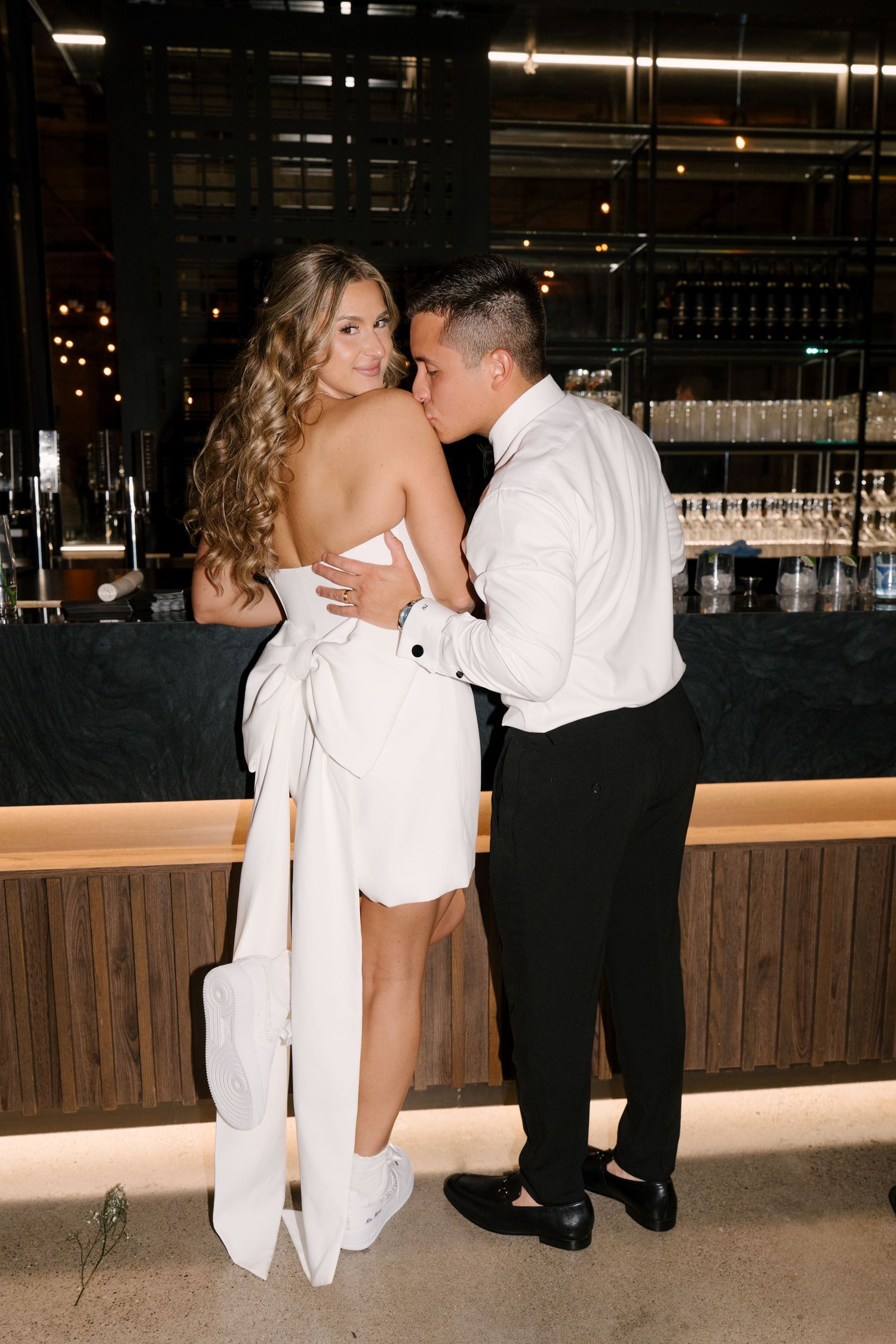 Bride in white strapless mini dress with bow and groom in white shirt share a tender moment at a modern bar in Tapestry Hall wedding venue.