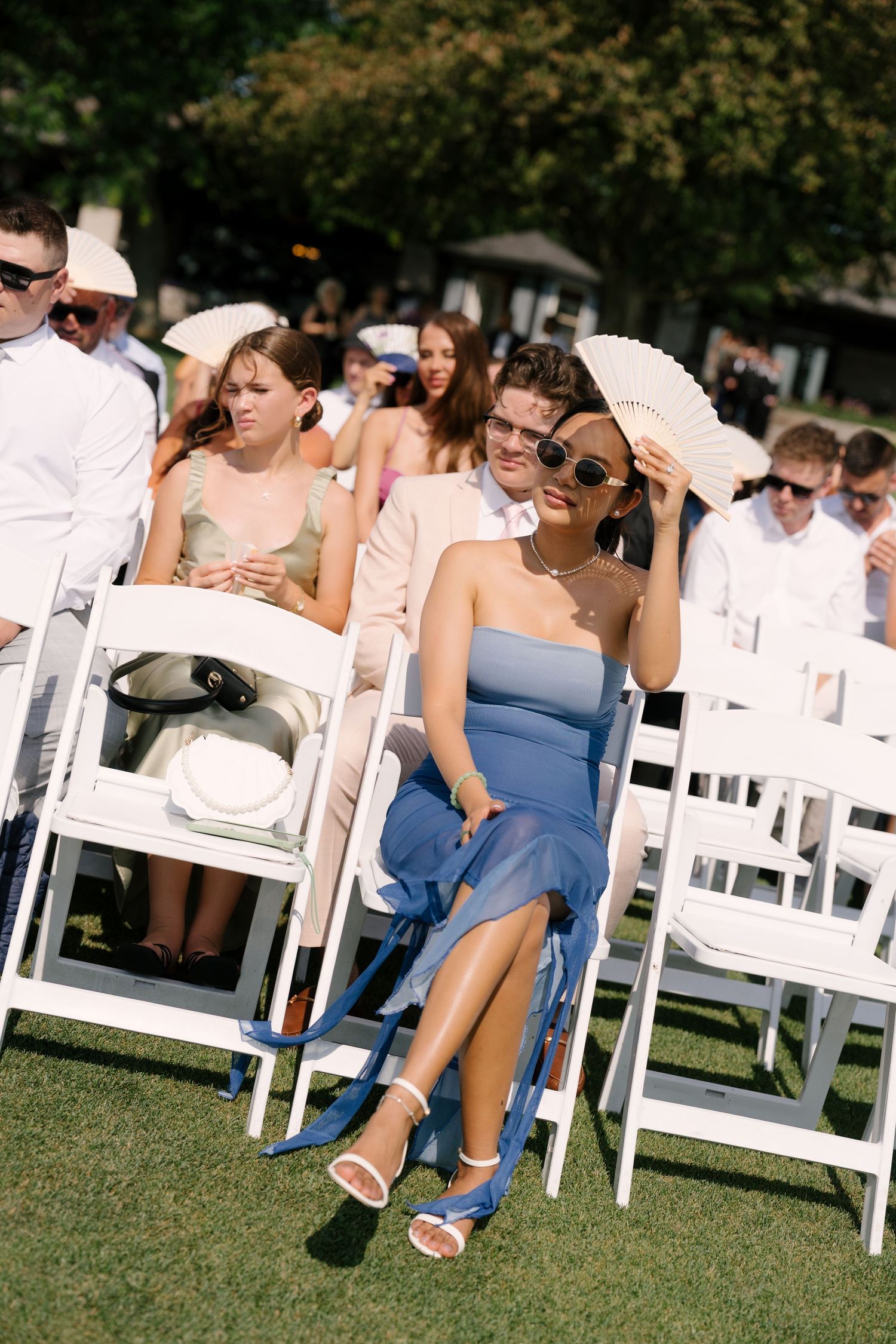 Wedding guests seated in white chairs outdoors, woman in blue strapless dress fans herself in bright sunlight.