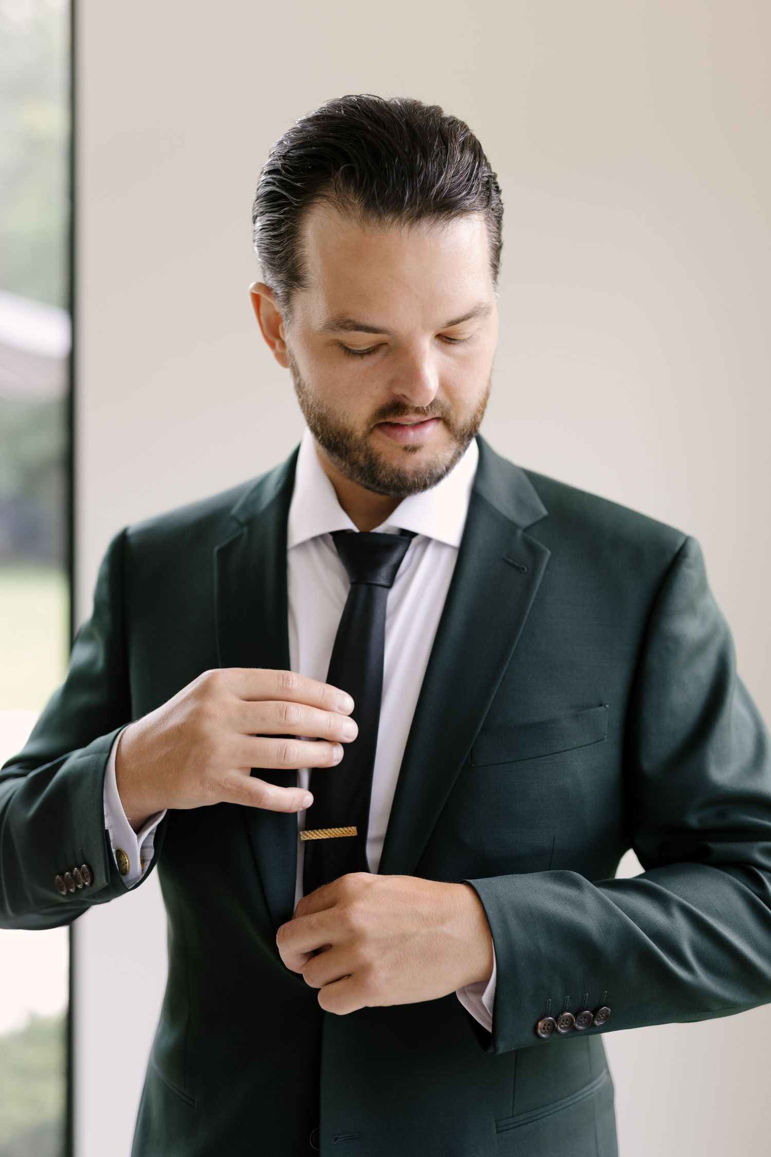 Dapper groom in dark green suit adjusting tie clip while getting ready at Roseville Estates wedding venue.