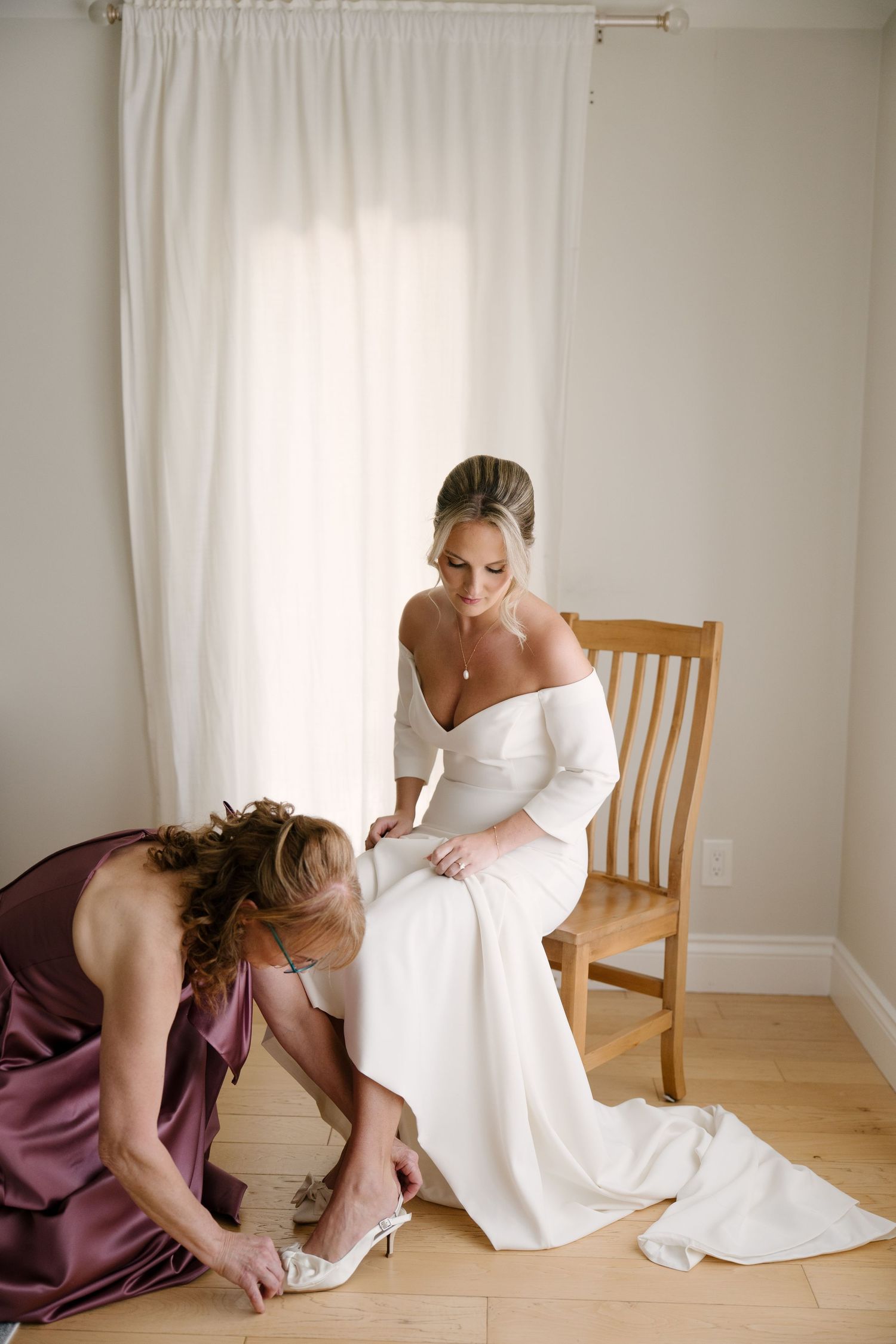 Bridesmaid in purple satin dress helping bride with her heels while seated on a wooden chair in soft light.