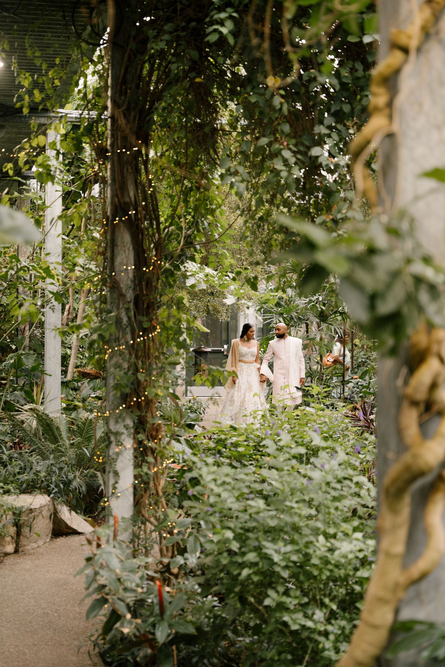 Wedding couple walks through a lush greenhouse garden surrounded by greenery and twinkling fairy lights.